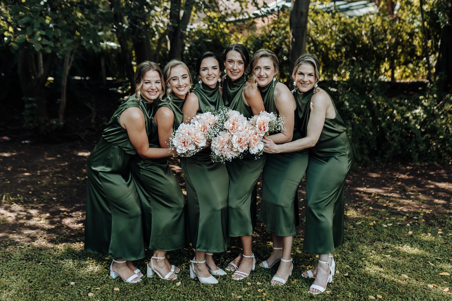 Group of bridesmaids in matching green dresses pose with bouquets outdoors.