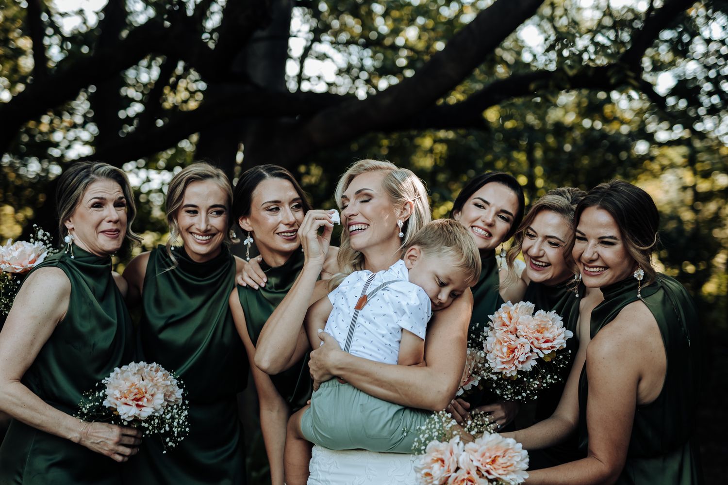 Bridal party in emerald dresses posing playfully with bouquets under tree canopy.