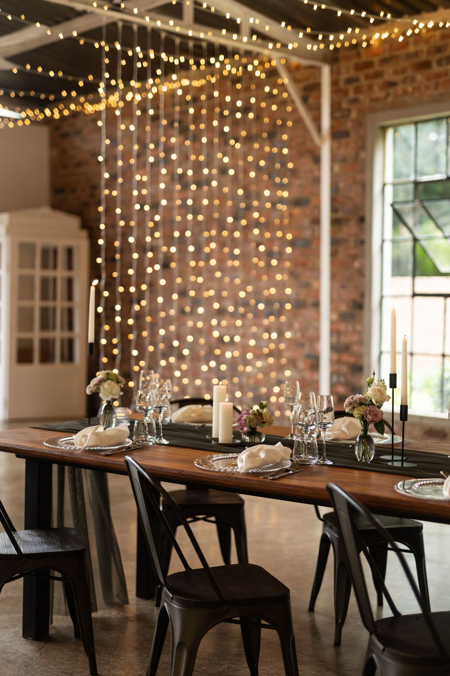 Industrial chic dining setup with exposed brick wall, string lights and modern black chairs.