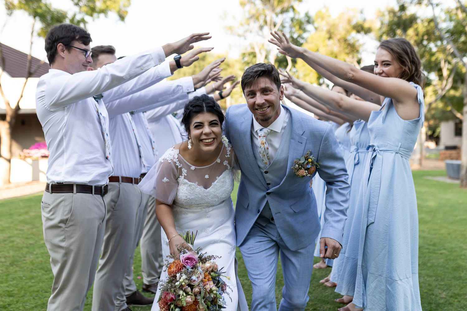 Wedding party forms tunnel of raised arms for couple to walk through on grassy lawn.
