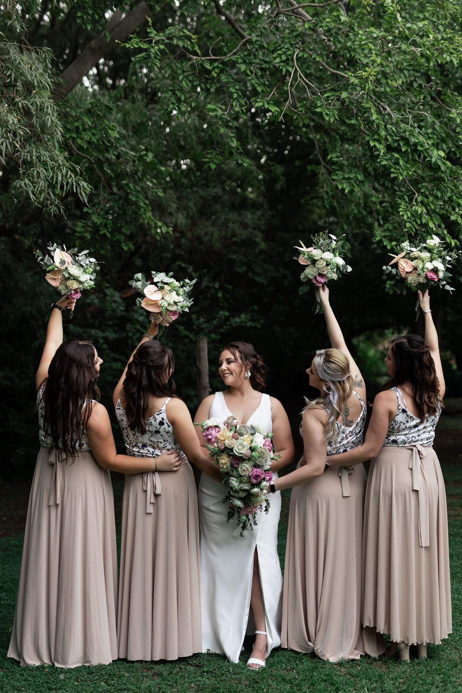 Wedding party in blush bridesmaid dresses raise floral bouquets while posing outdoors under green trees.