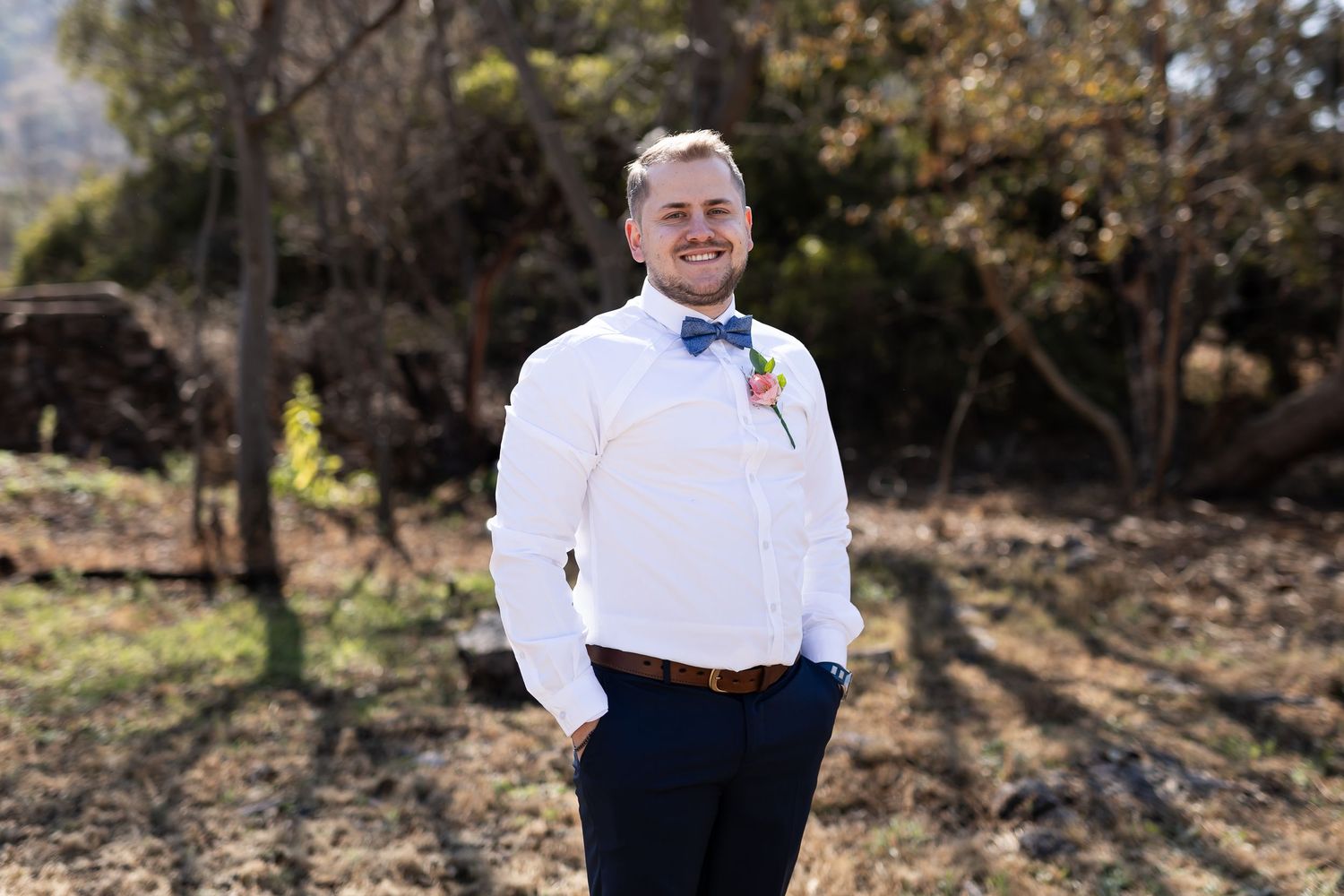A person in a formal white shirt and black bowtie poses outdoors against a blurred natural backdrop during a photo session.