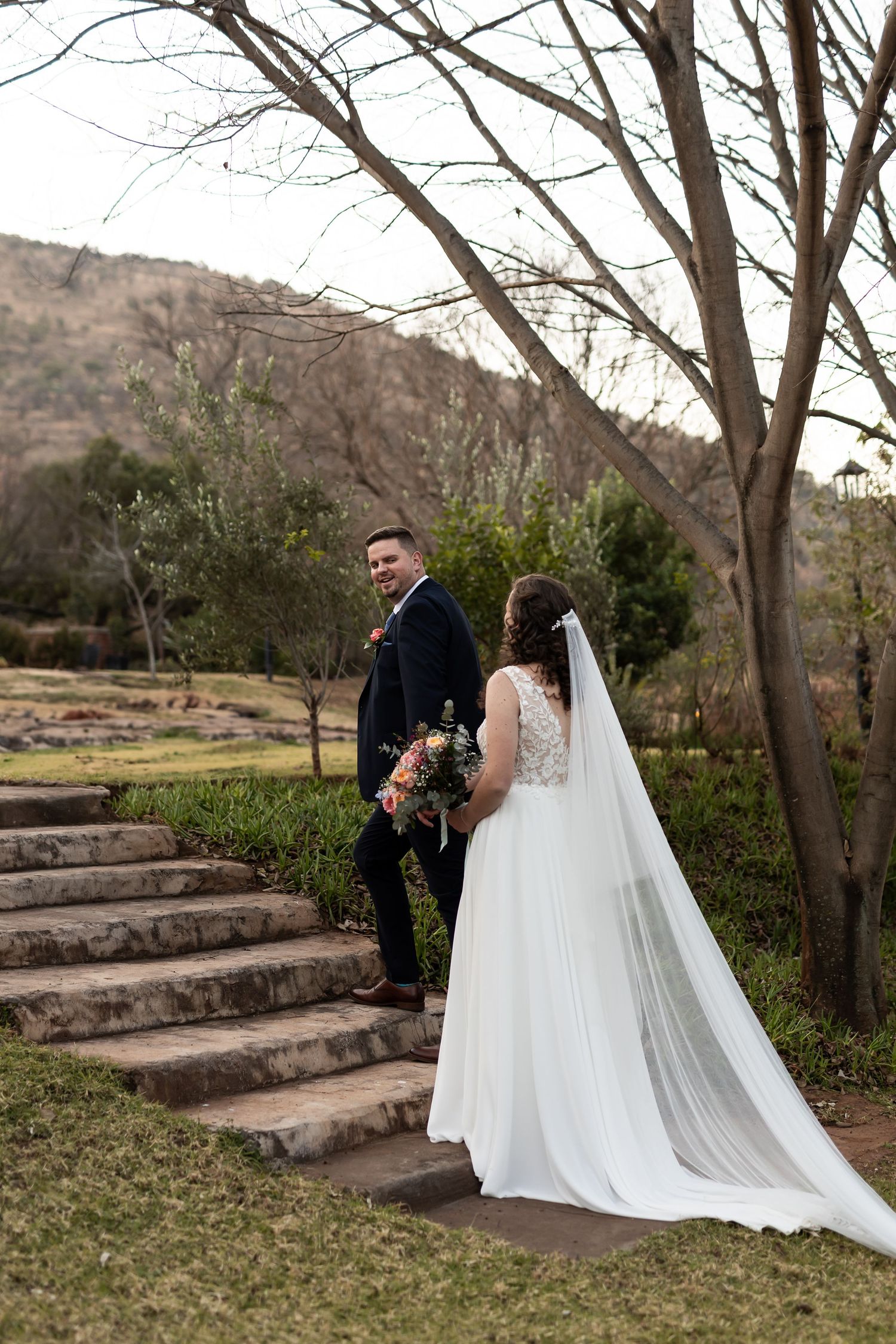 Newlywed couple stands on rustic stone steps in a garden setting with mountains in the background.