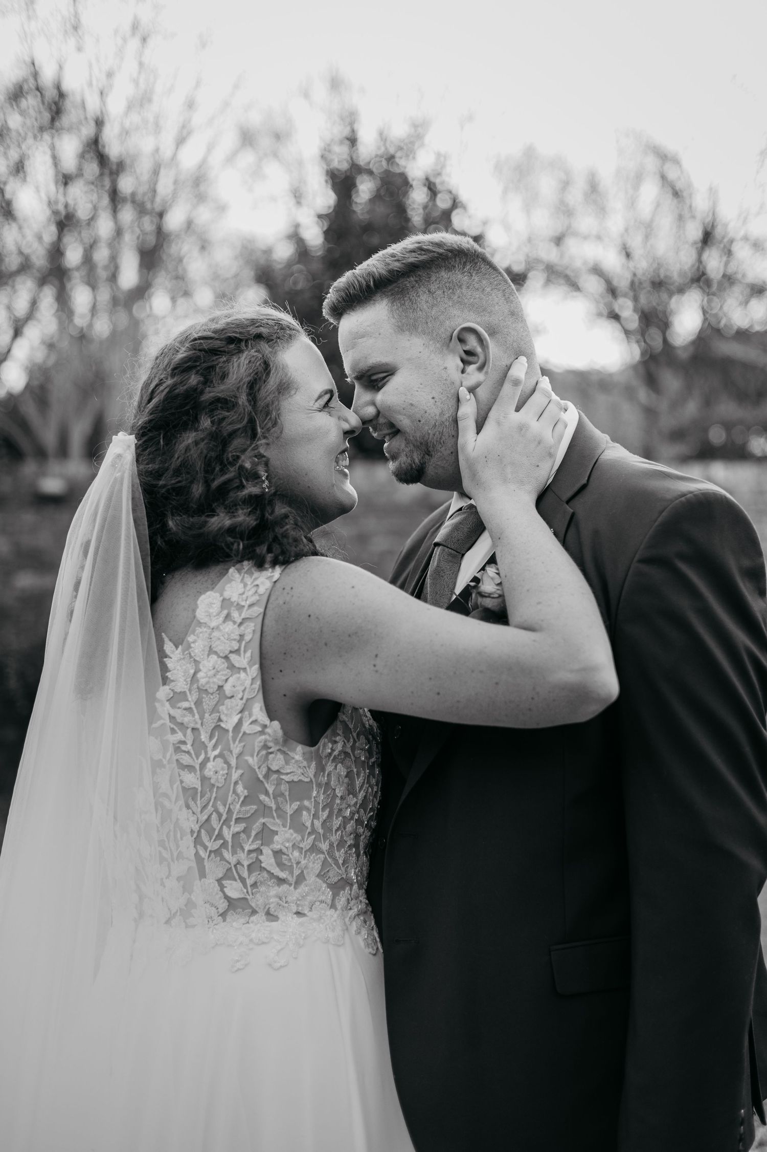 Intimate black and white wedding portrait showing tender moment between newlyweds in lace gown and dark suit outdoors.