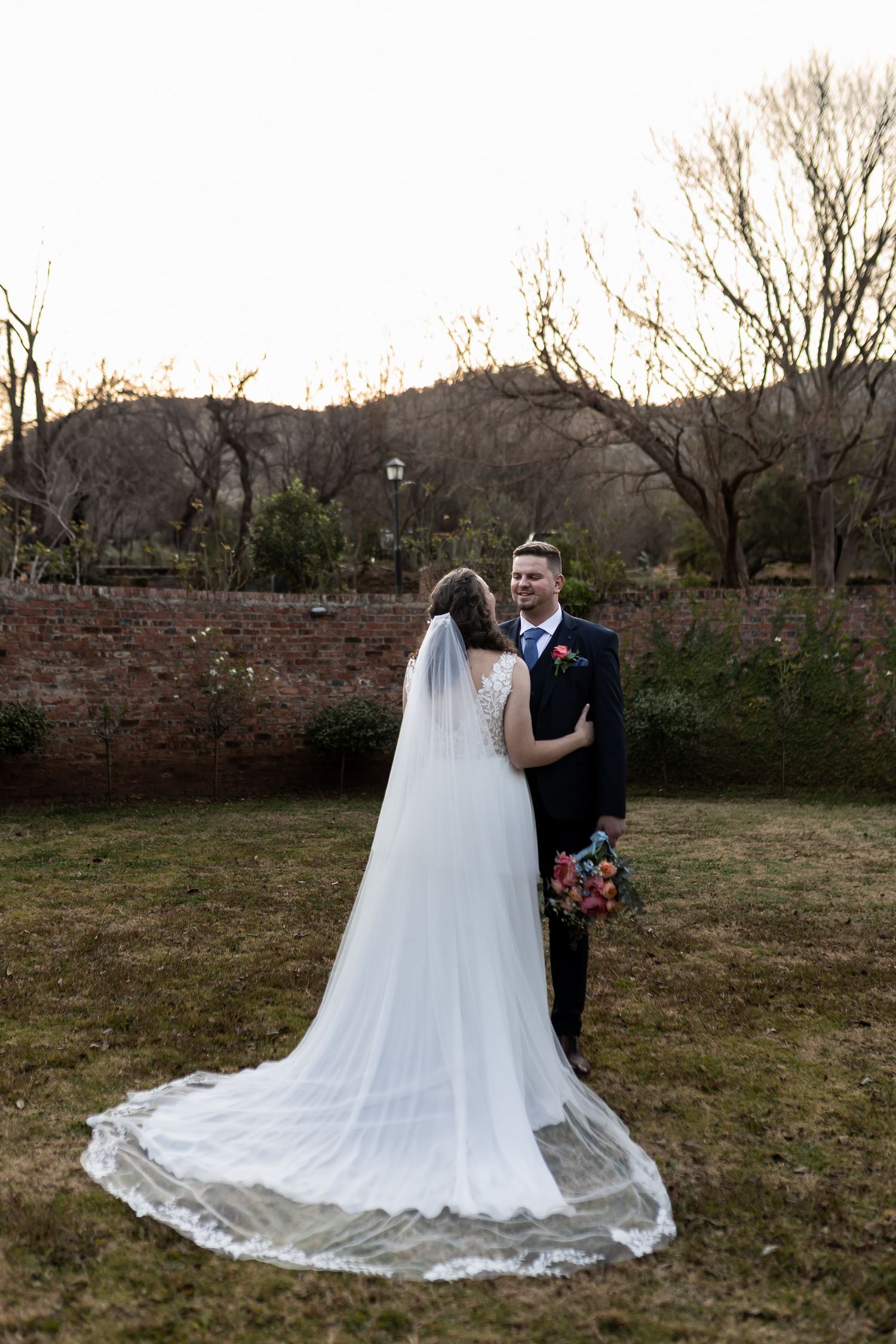 Couple poses in formal wedding attire in front of stone wall and barren trees during sunset with flowing white veil.