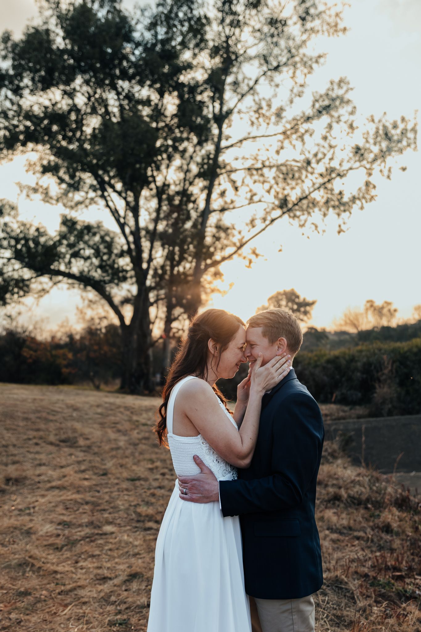 A romantic couple embraces during sunset in a field with autumn trees in the background.