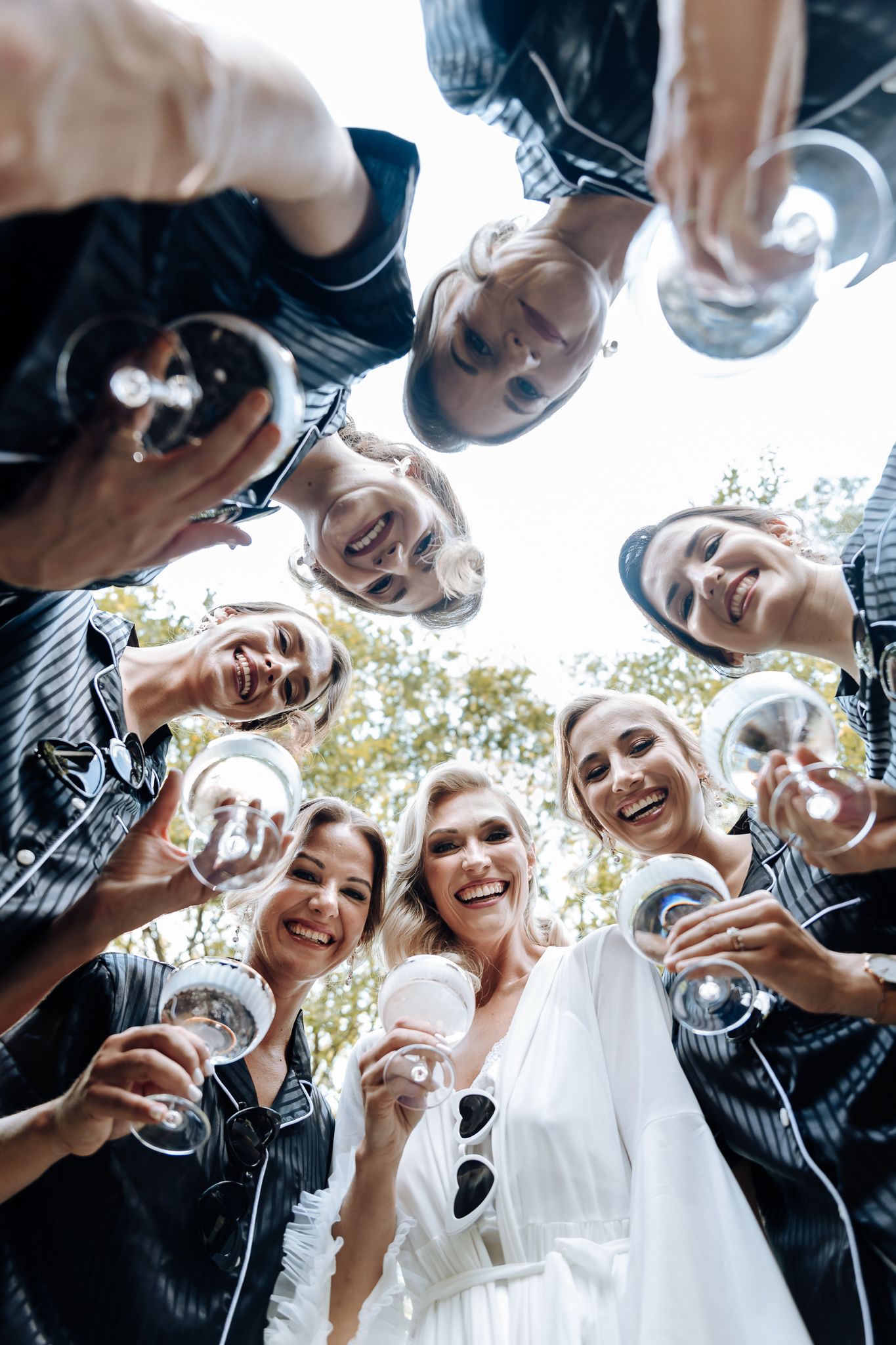 Wedding party gathered in a circle from below toasting with champagne glasses against a sunny sky.