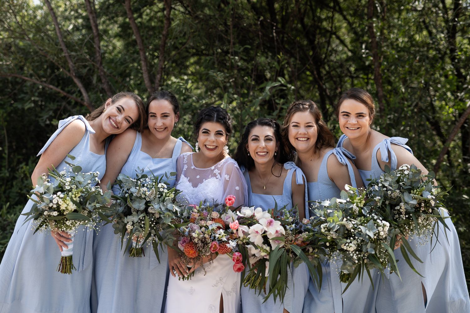 A bridal party in powder blue dresses poses outdoors while holding floral bouquets.
