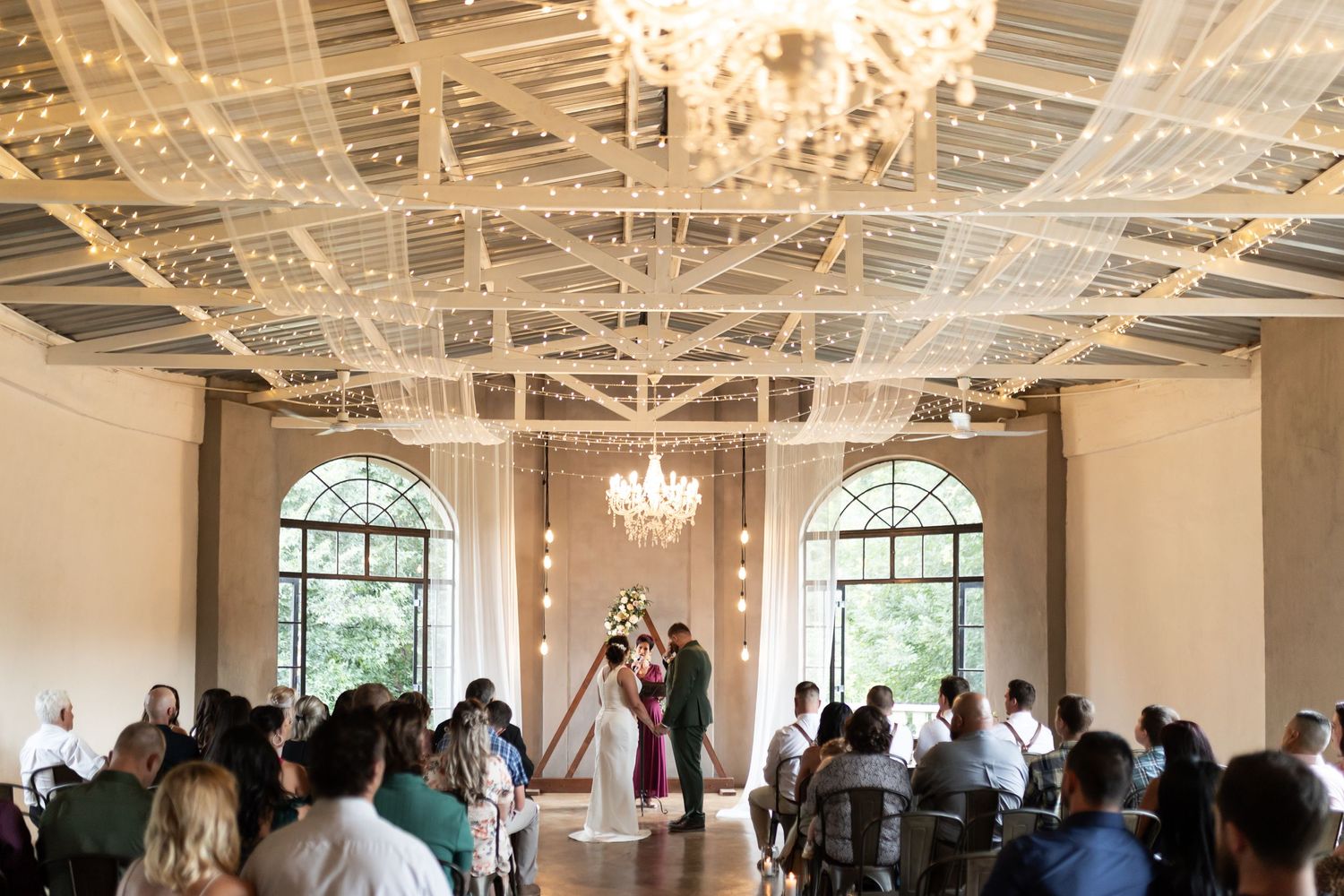 A wide shot of a wedding ceremony in an elegant room with chandeliers and large arched windows.