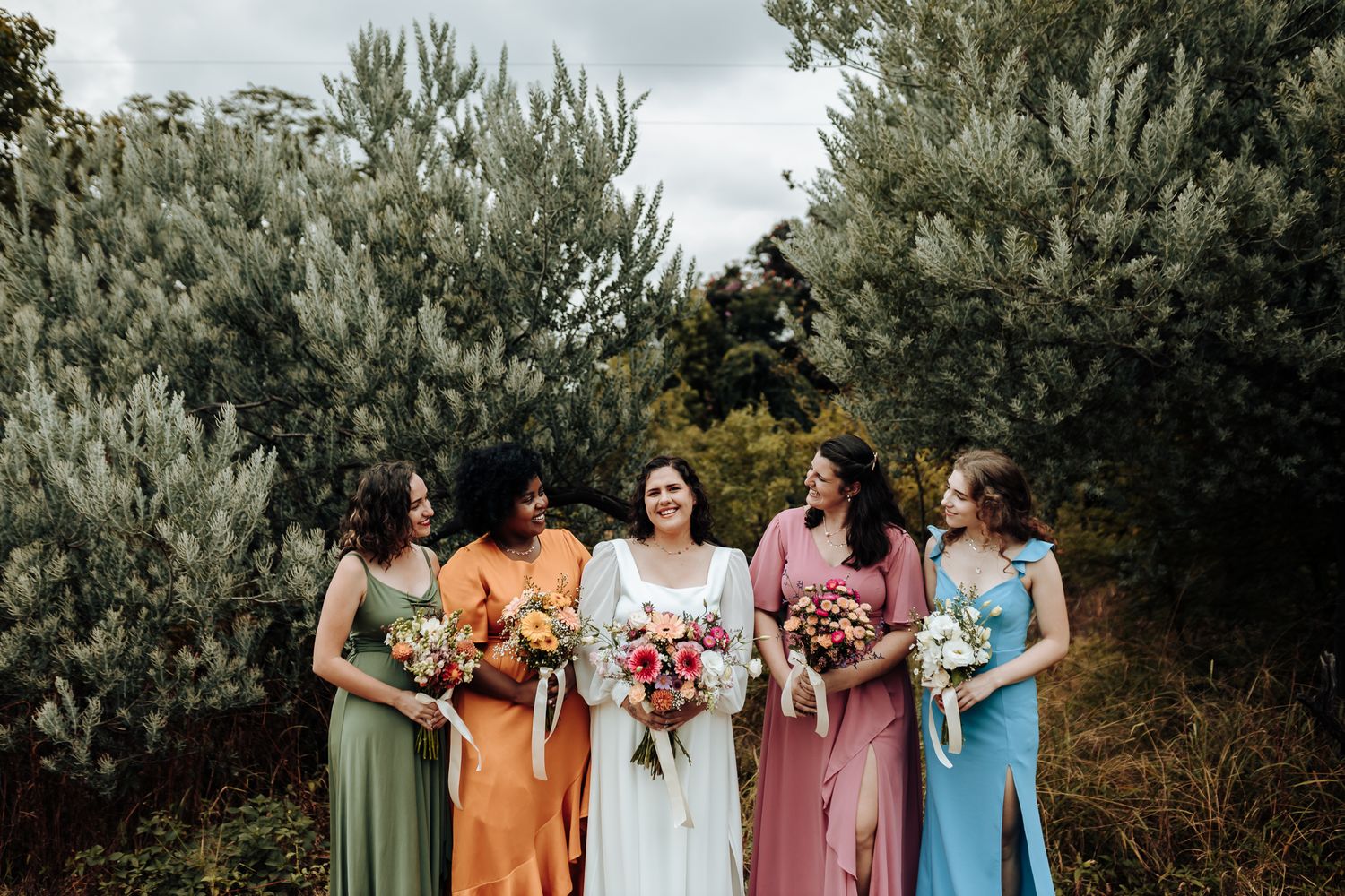 Wedding party in colorful dresses ranging from sage green to orange to pink stand together holding bouquets.