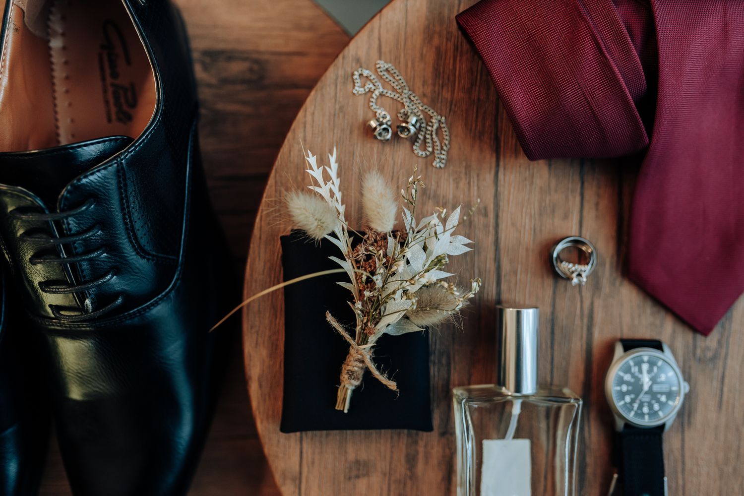 Wedding accessories including shoes, tie, watch and dried flowers arranged on wooden surface.