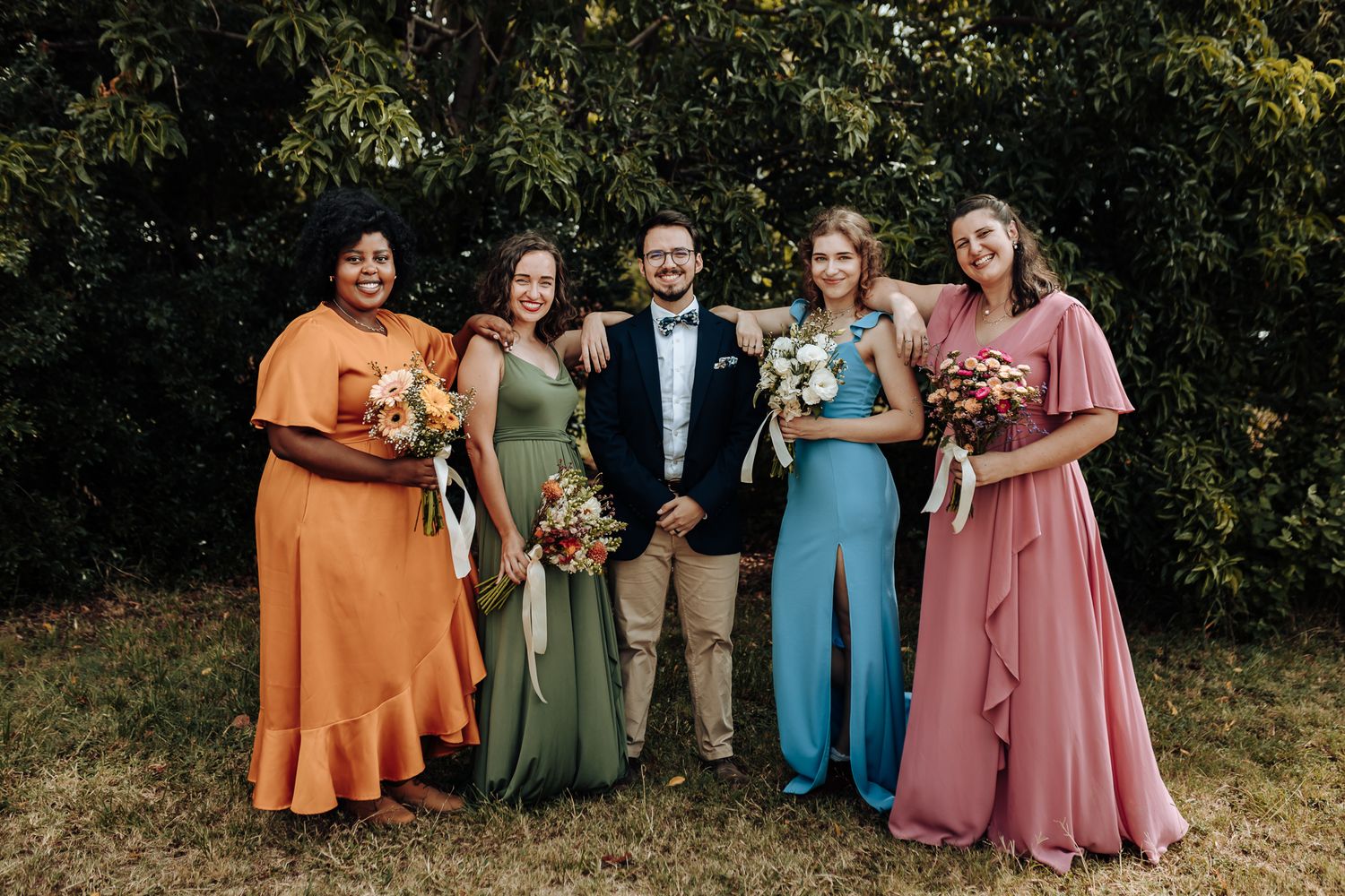 Wedding party in colorful dresses posing with someone in formal black attire outdoors.