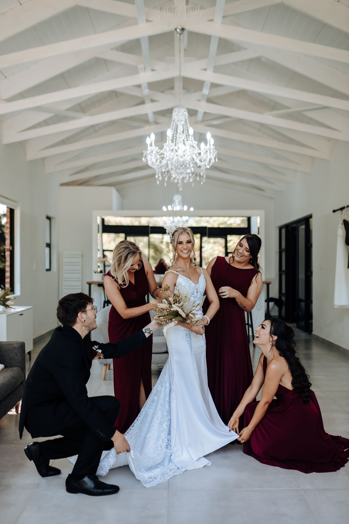 Bridal party in burgundy dresses gather around bride in elegant white venue with chandelier.