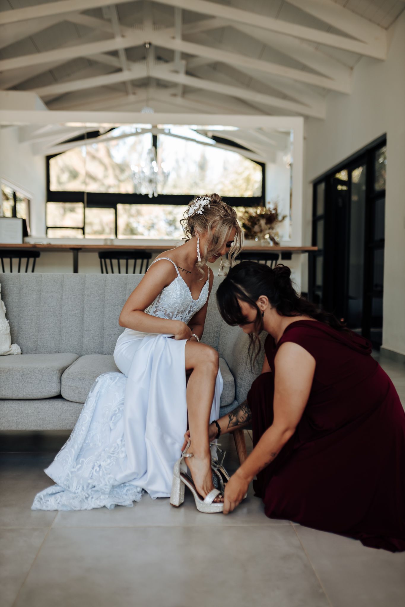 Helper in burgundy dress assists with wedding shoe fitting on gray couch in modern white room.