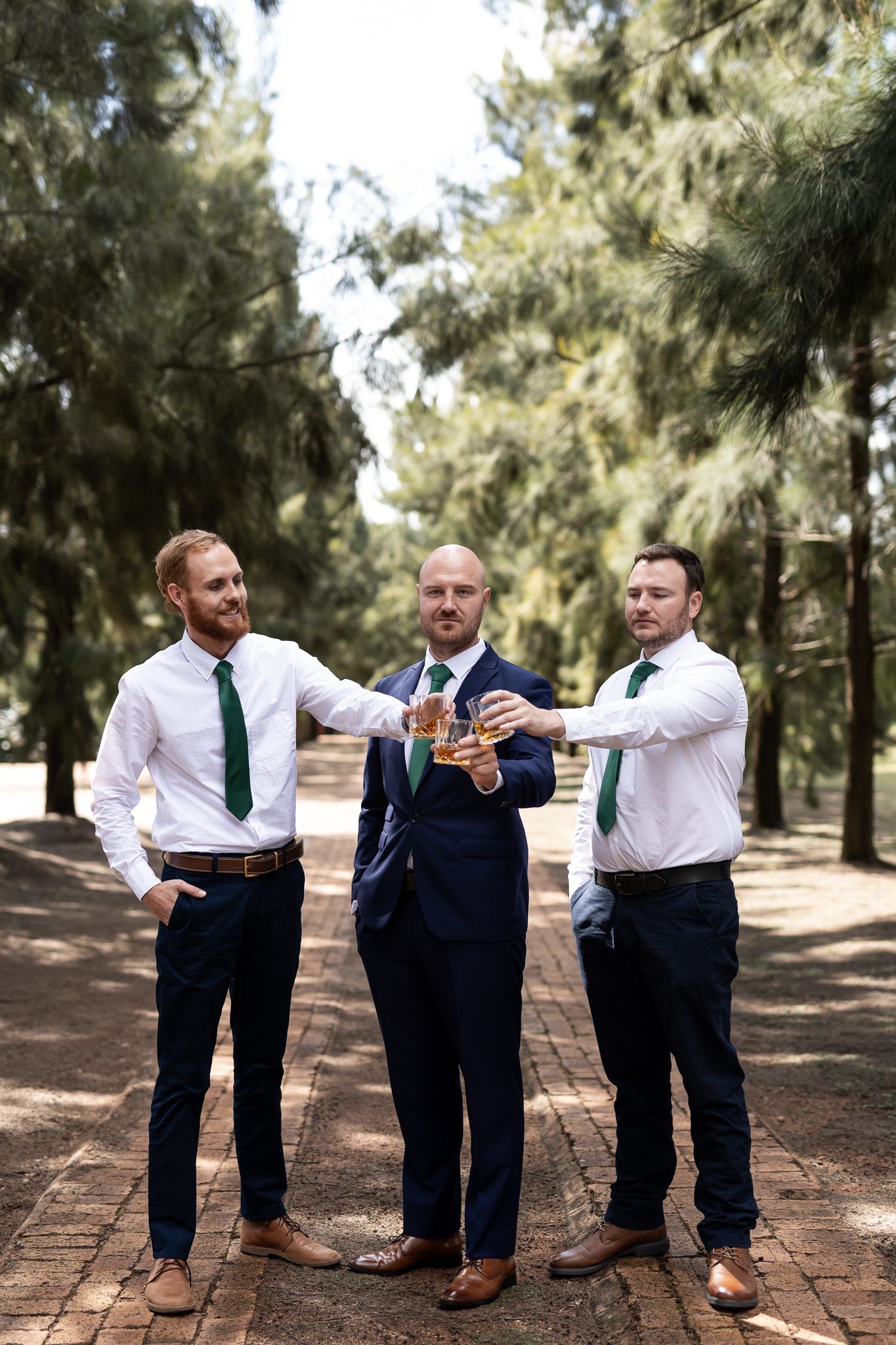 Three groomsmen in coordinated attire posing playfully on brick walkway in pine forest.