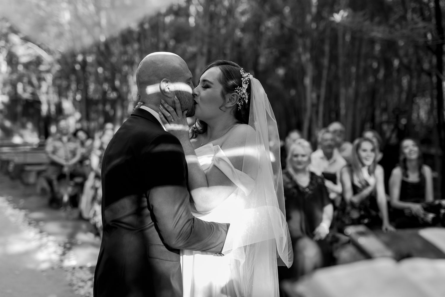 A tender moment captured during a woodland wedding ceremony with guests watching.