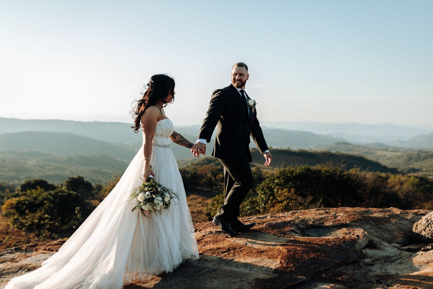 Wedding couple walks hand in hand on mountain summit with flowing white dress catching the breeze.