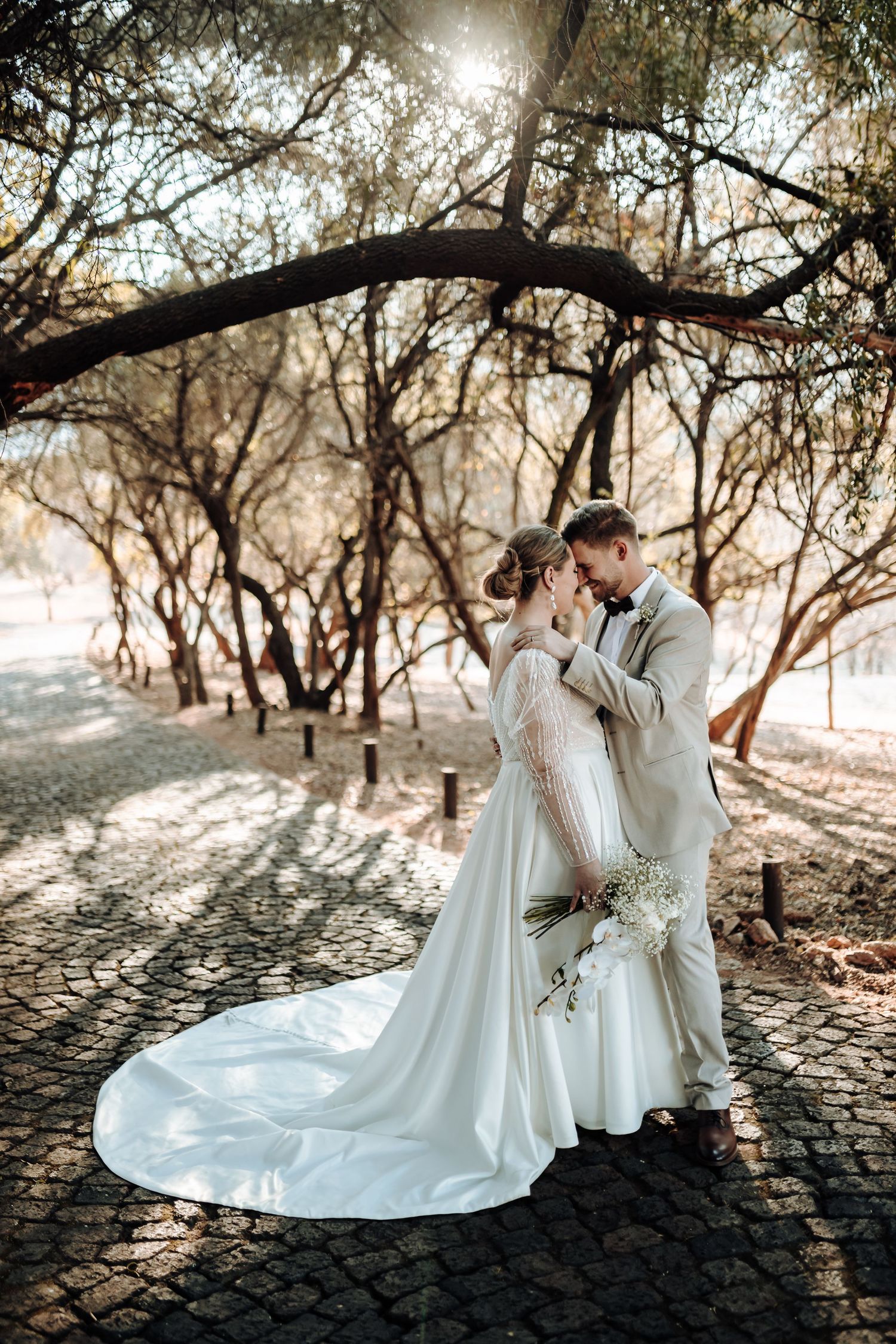 Romantic wedding couple embrace under arched trees on a stone path in warm sunlit forest setting.