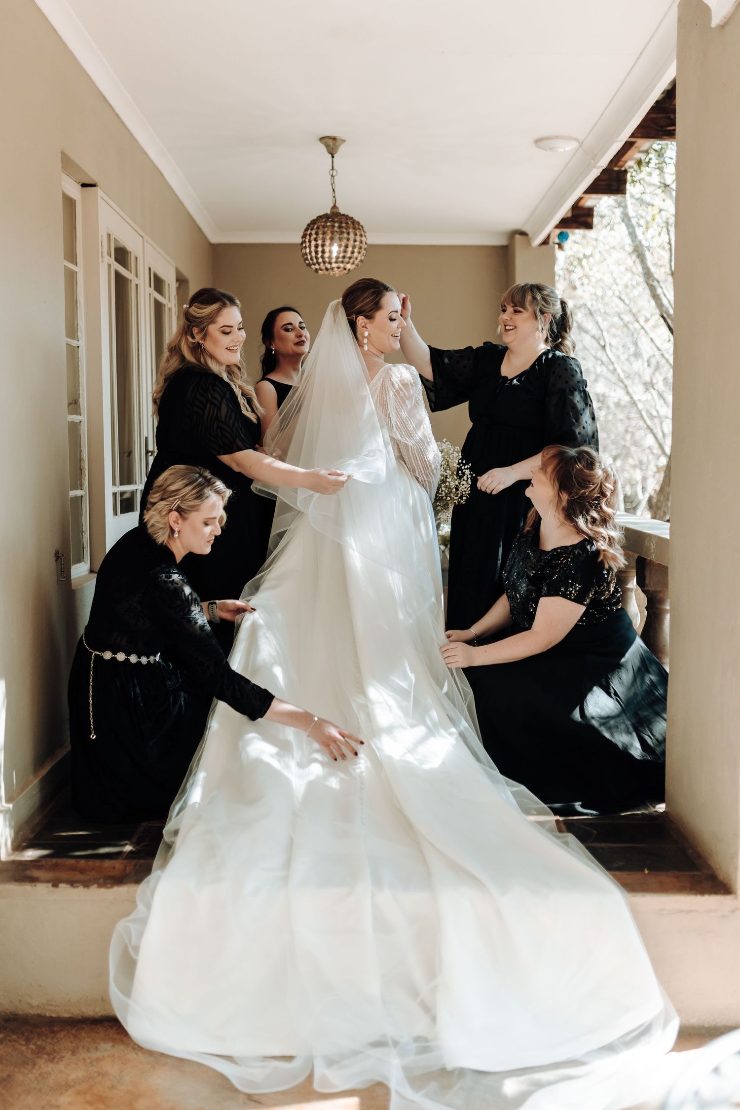 Bridesmaids help arrange bride's wedding dress train in elegant hallway.