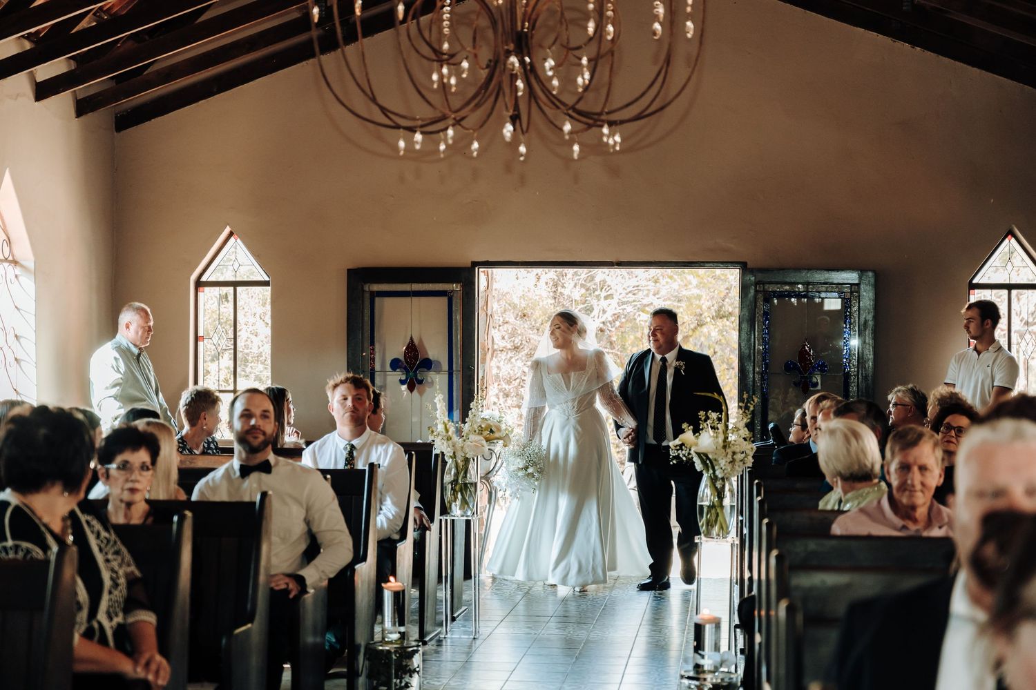 Wedding ceremony procession in rustic chapel with chandelier and wooden beam ceiling.