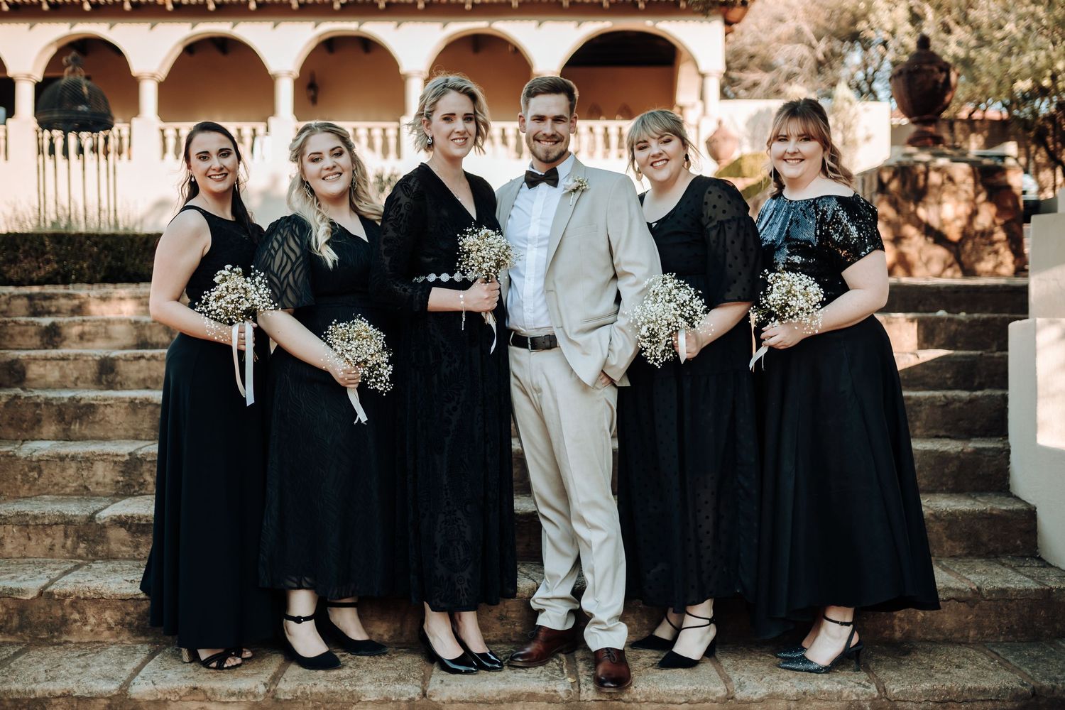 Wedding party in black and white attire poses on stone steps of Spanish colonial building.