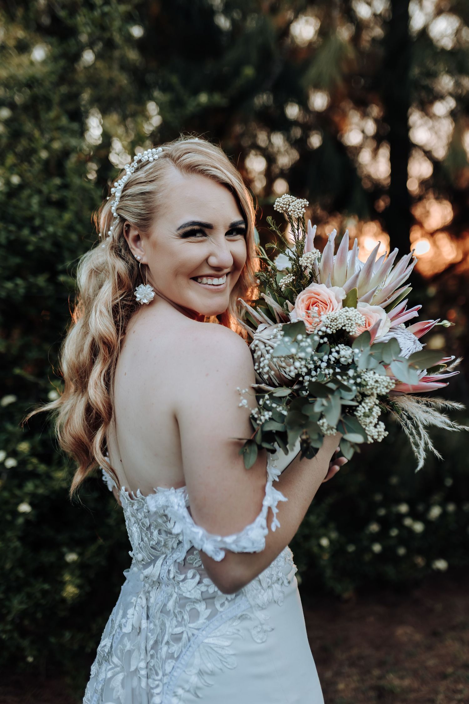 Bride in lace wedding gown turns to show bouquet featuring protea and greenery at sunset.