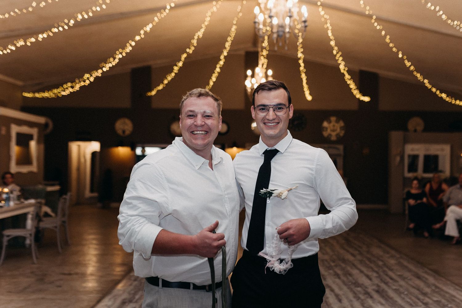 Two guests in white shirts smile at the camera during a wedding reception.