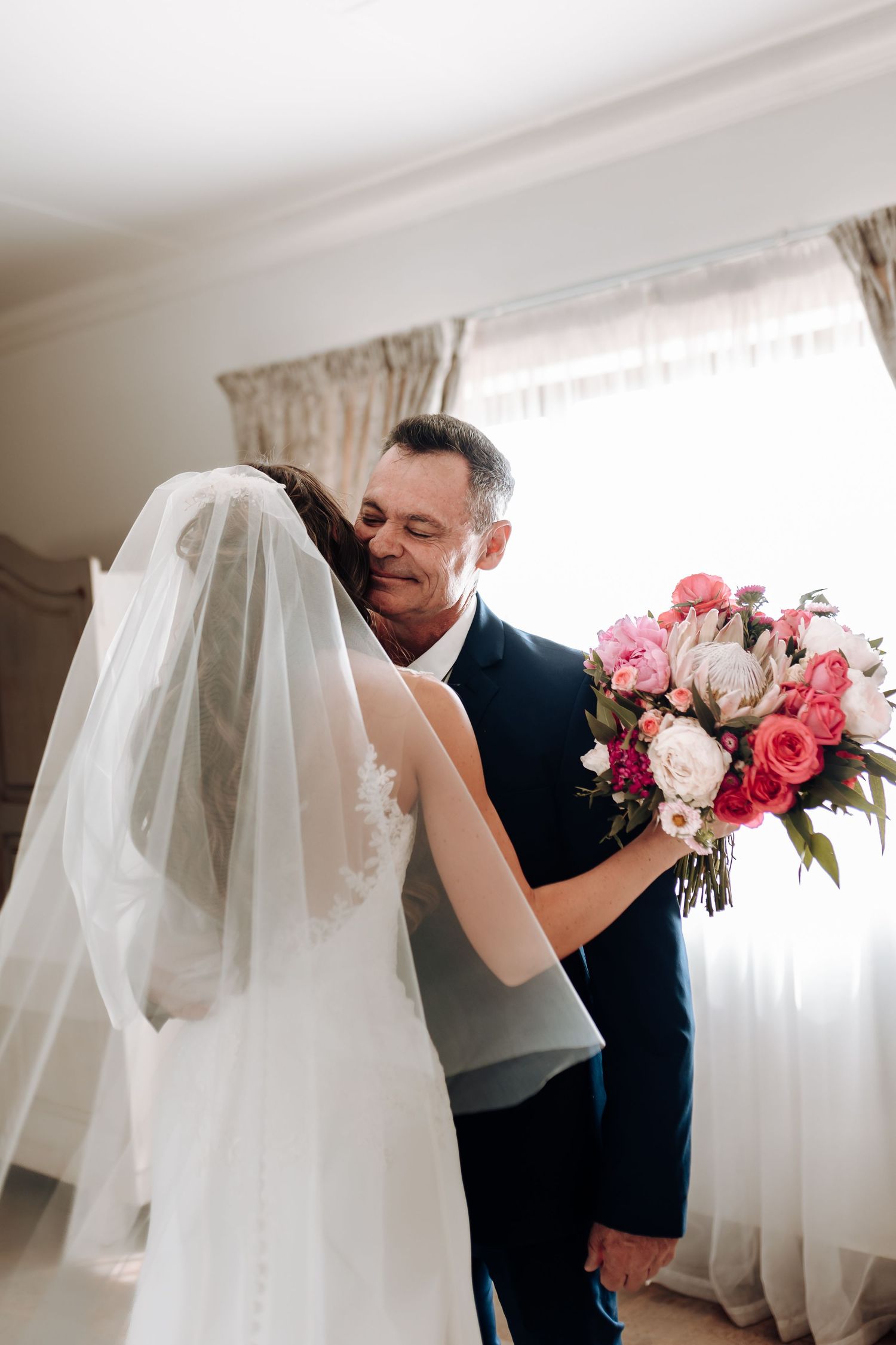 Emotional first look between bride and groom with pink bouquet by window.