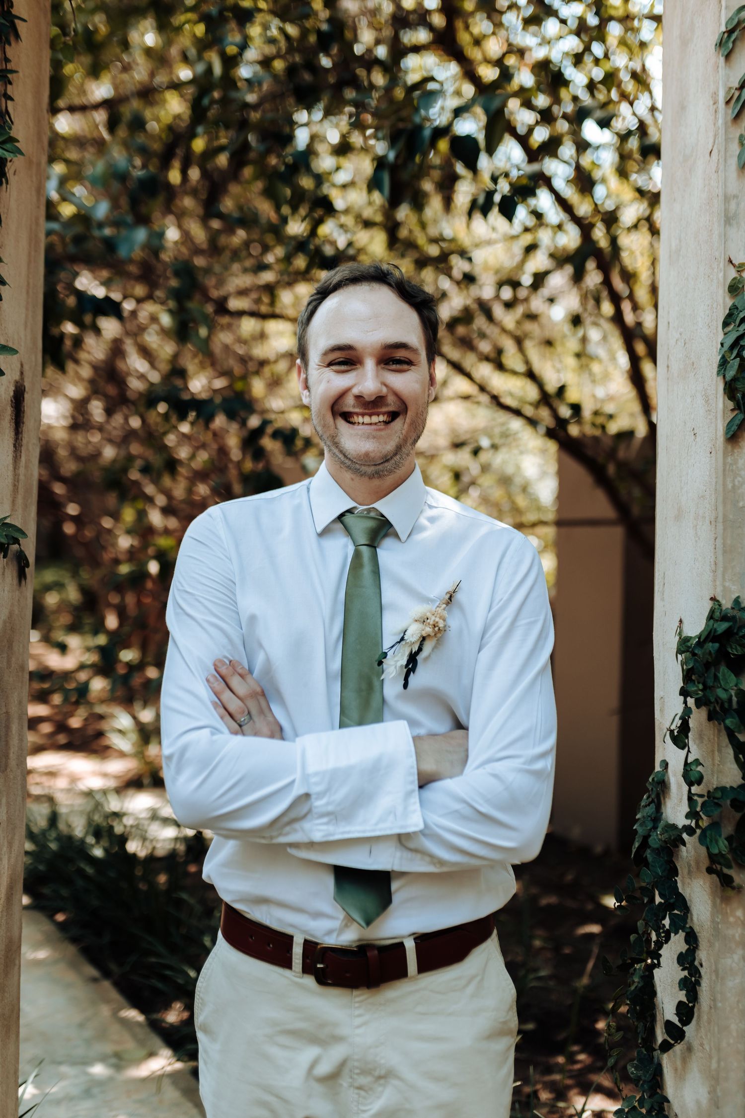 Person in white shirt and green tie smiles with arms crossed in front of vine-covered wall.
