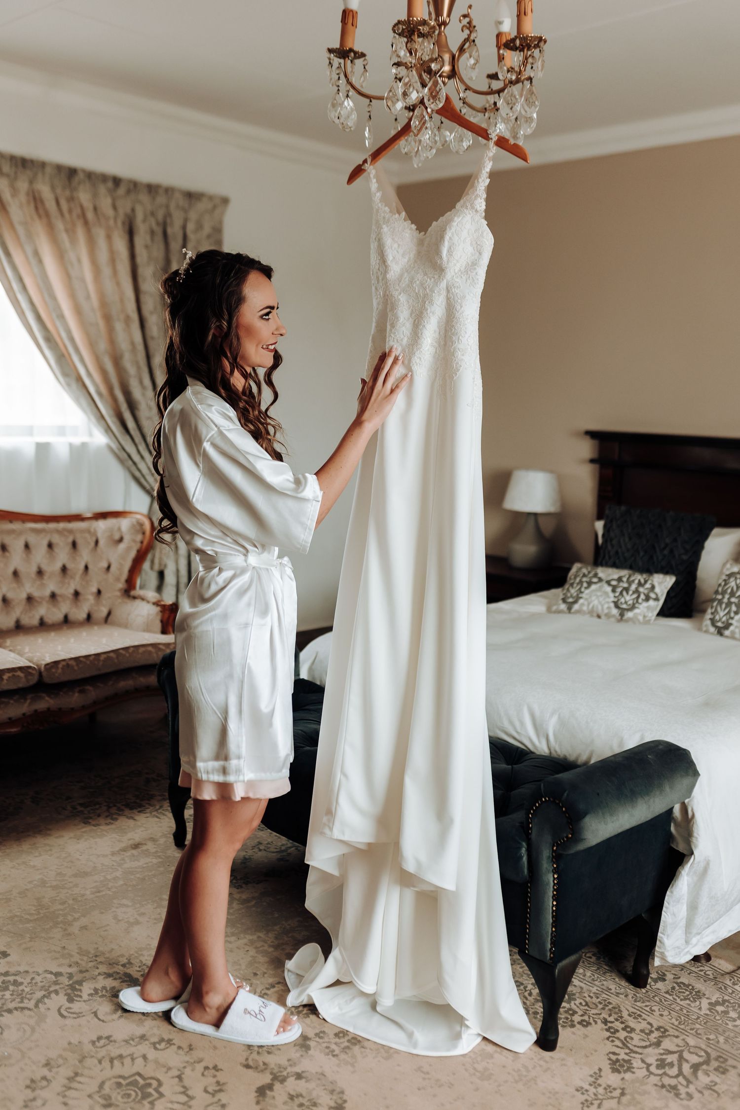 Getting ready scene with white wedding gown hanging from chandelier in elegant bedroom.
