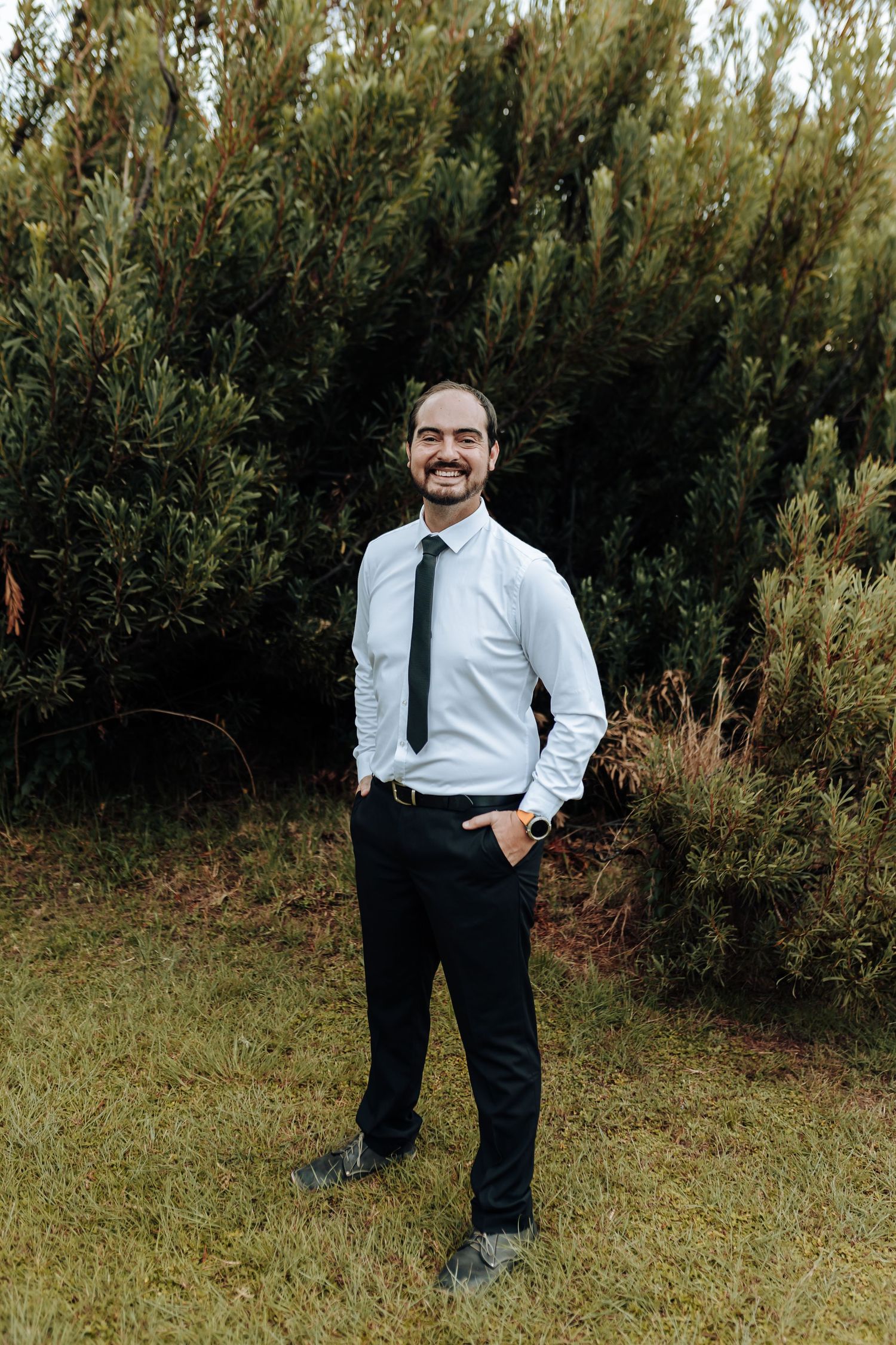 A smiling man in white dress shirt and tie poses casually against a backdrop of green trees.