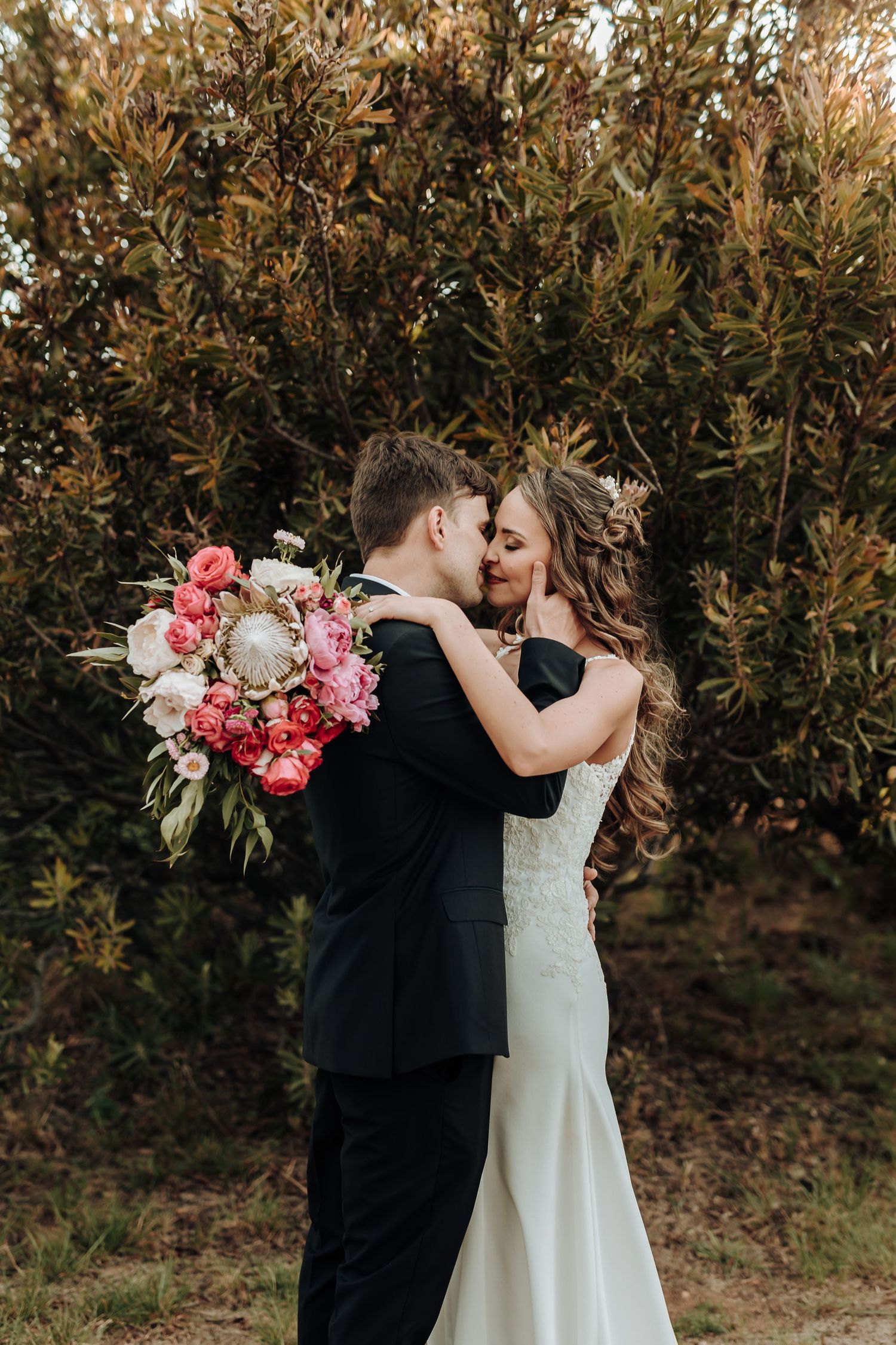Newlyweds share an intimate moment with pink floral bouquet against foliage.