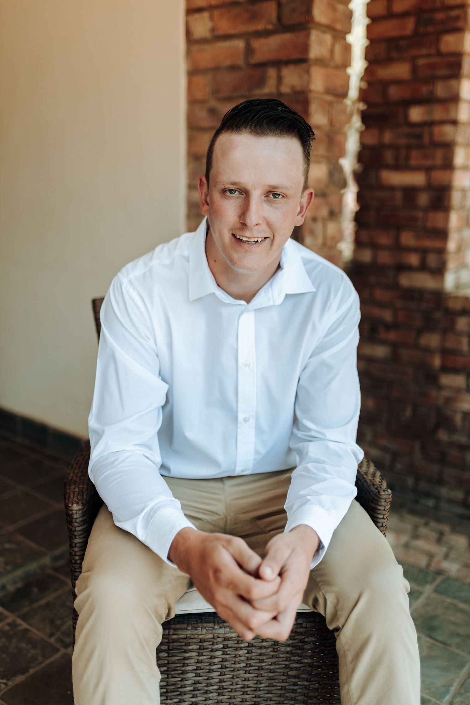 A seated figure in crisp white shirt and khaki pants against a brick wall backdrop.