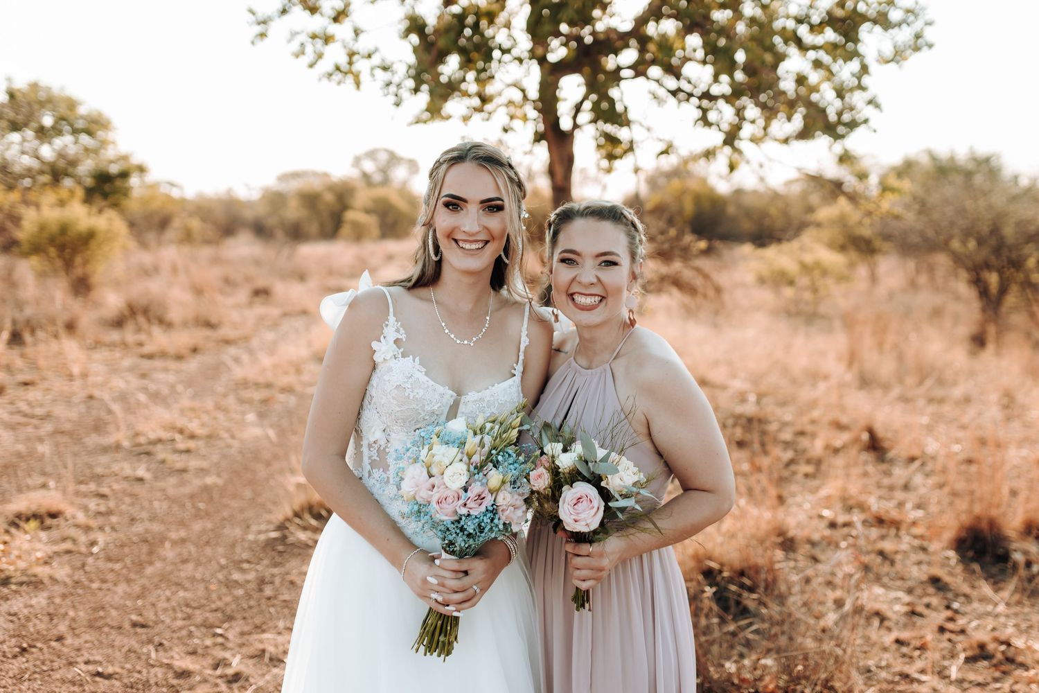 Wedding party members in light pink and white dresses holding floral bouquets against a warm rustic backdrop.