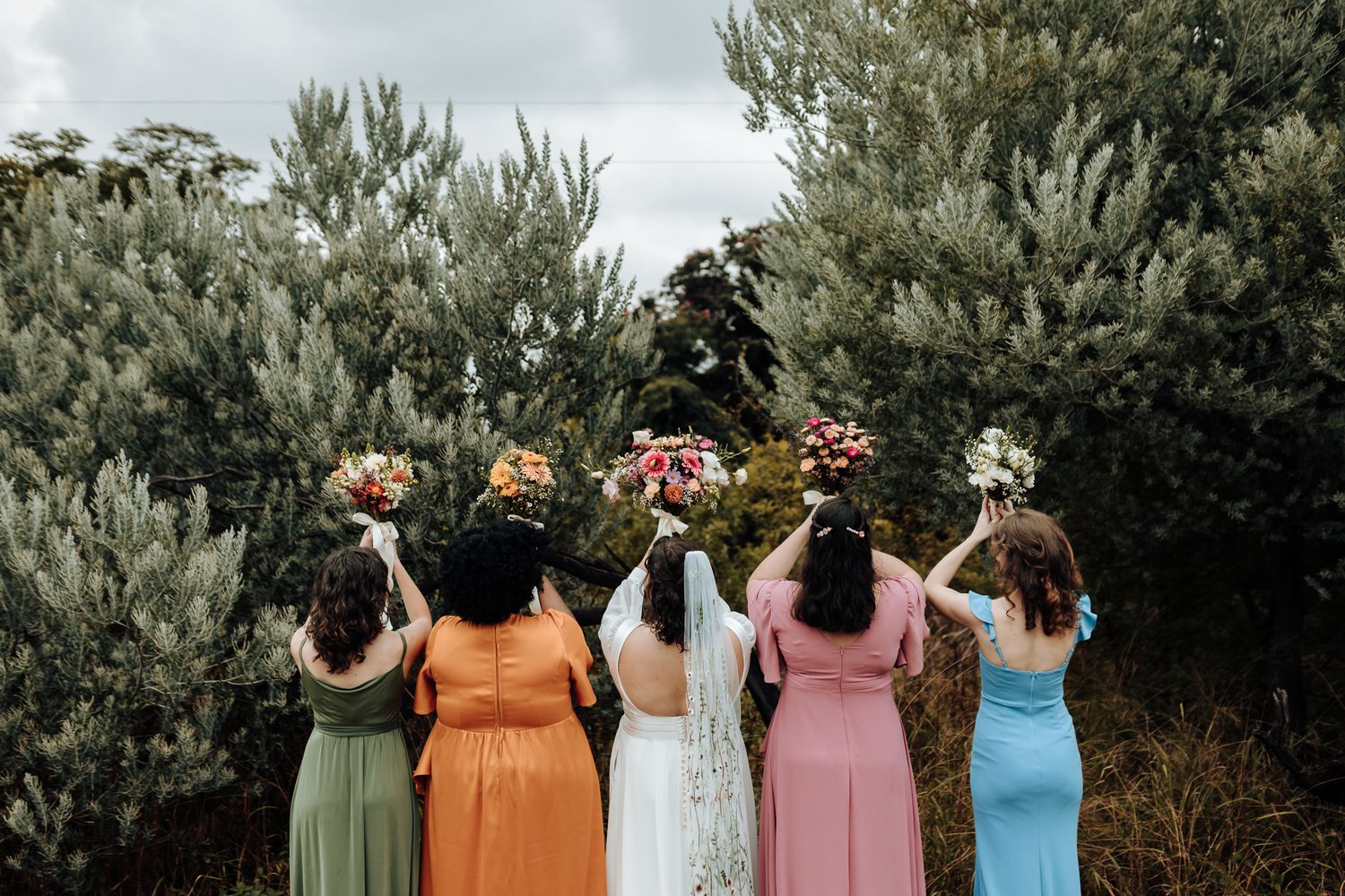 Back view of bridal party in colorful dresses holding bouquets up against a cloudy sky.