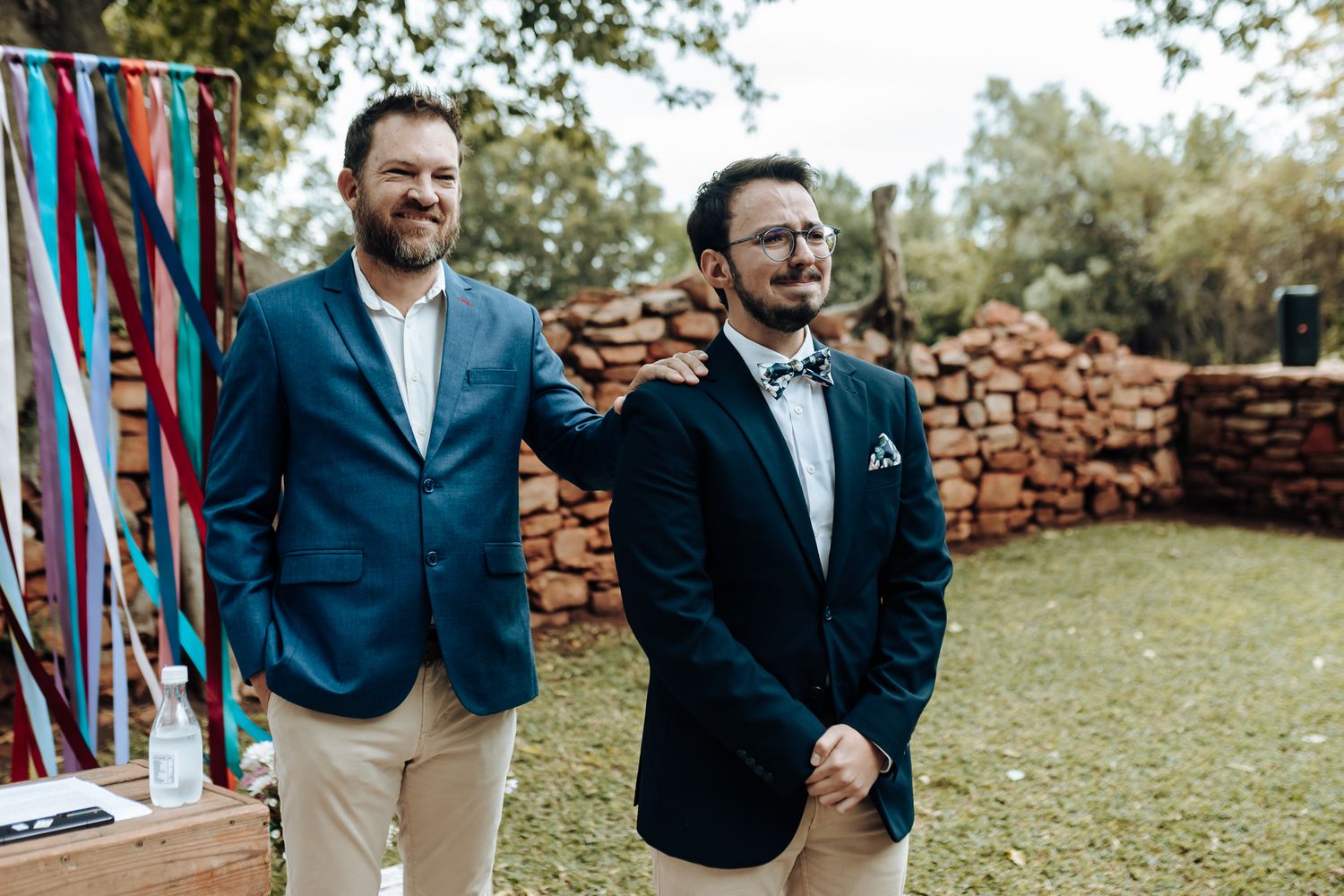 Two men in formal suits stand during outdoor wedding ceremony near colorful ribbon decor.