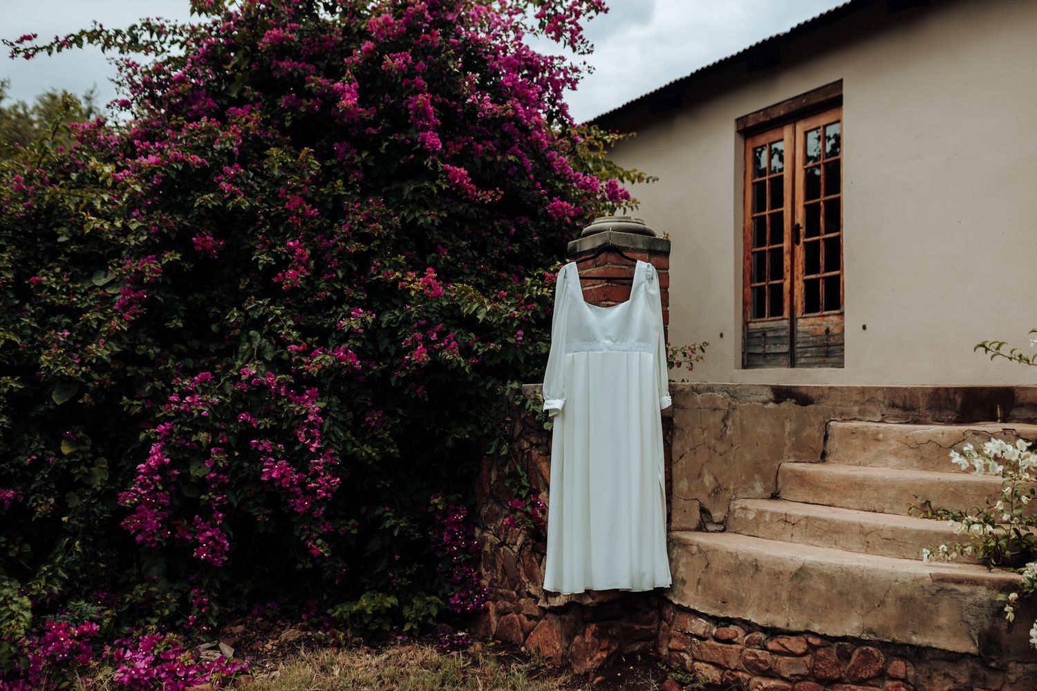 White dress hanging on display against a stone house with vibrant pink bougainvillea flowers.