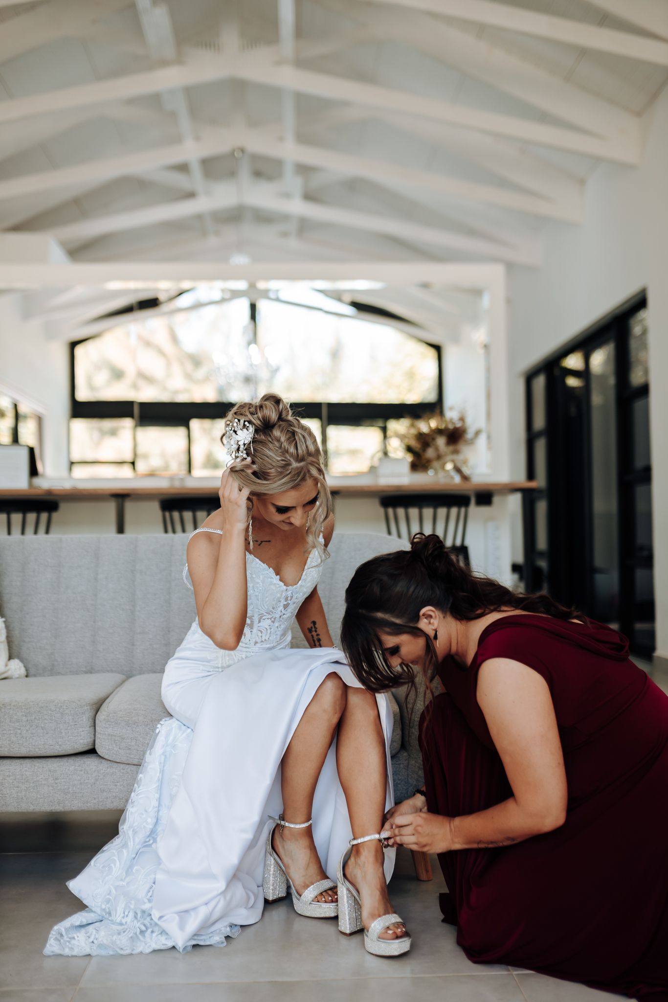 Helper in burgundy dress assists with wedding shoe adjustments in bright modern setting.
