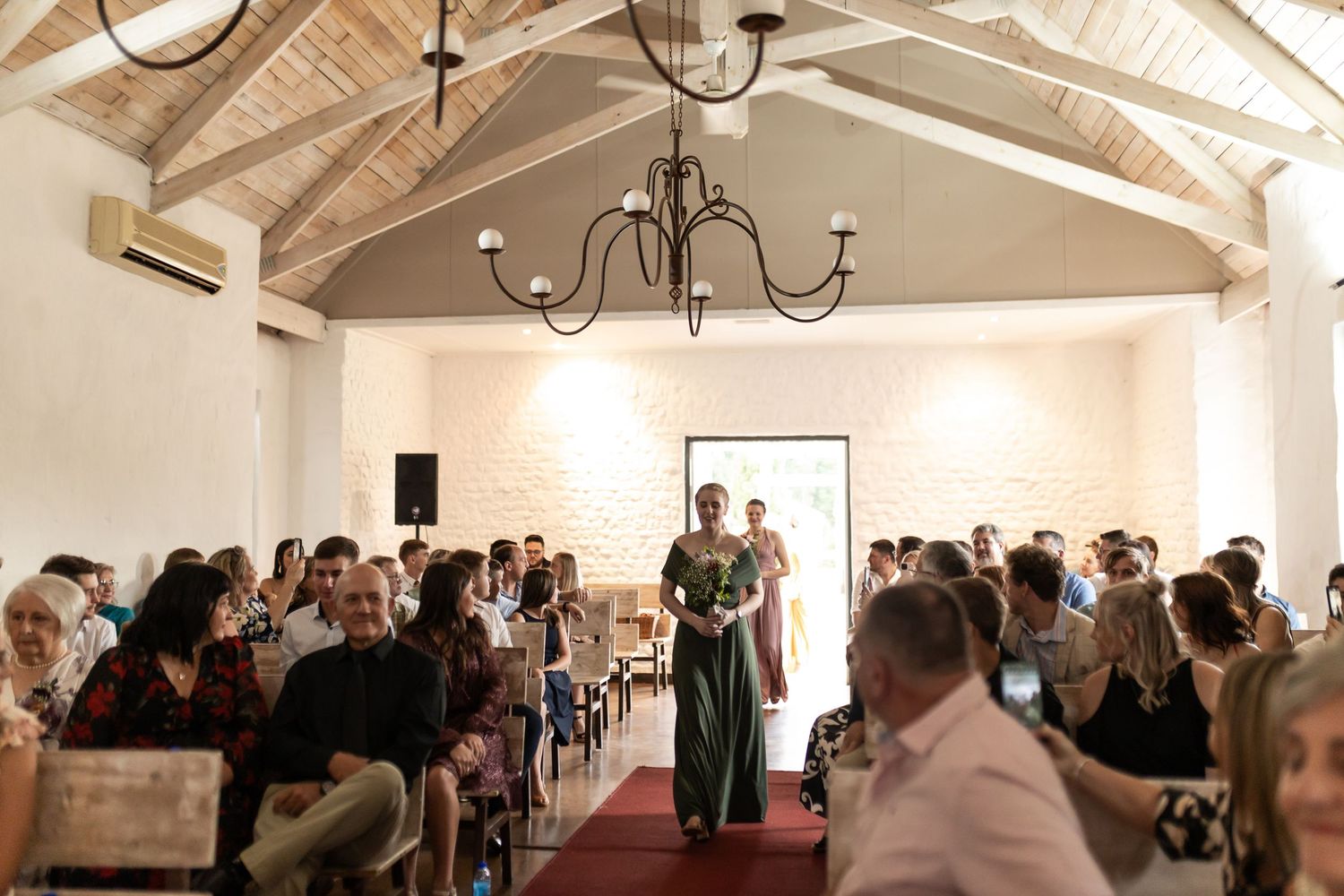 Guests seated in a chapel with vaulted ceilings and chandeliers watch as attendants walk down the red-carpeted aisle.