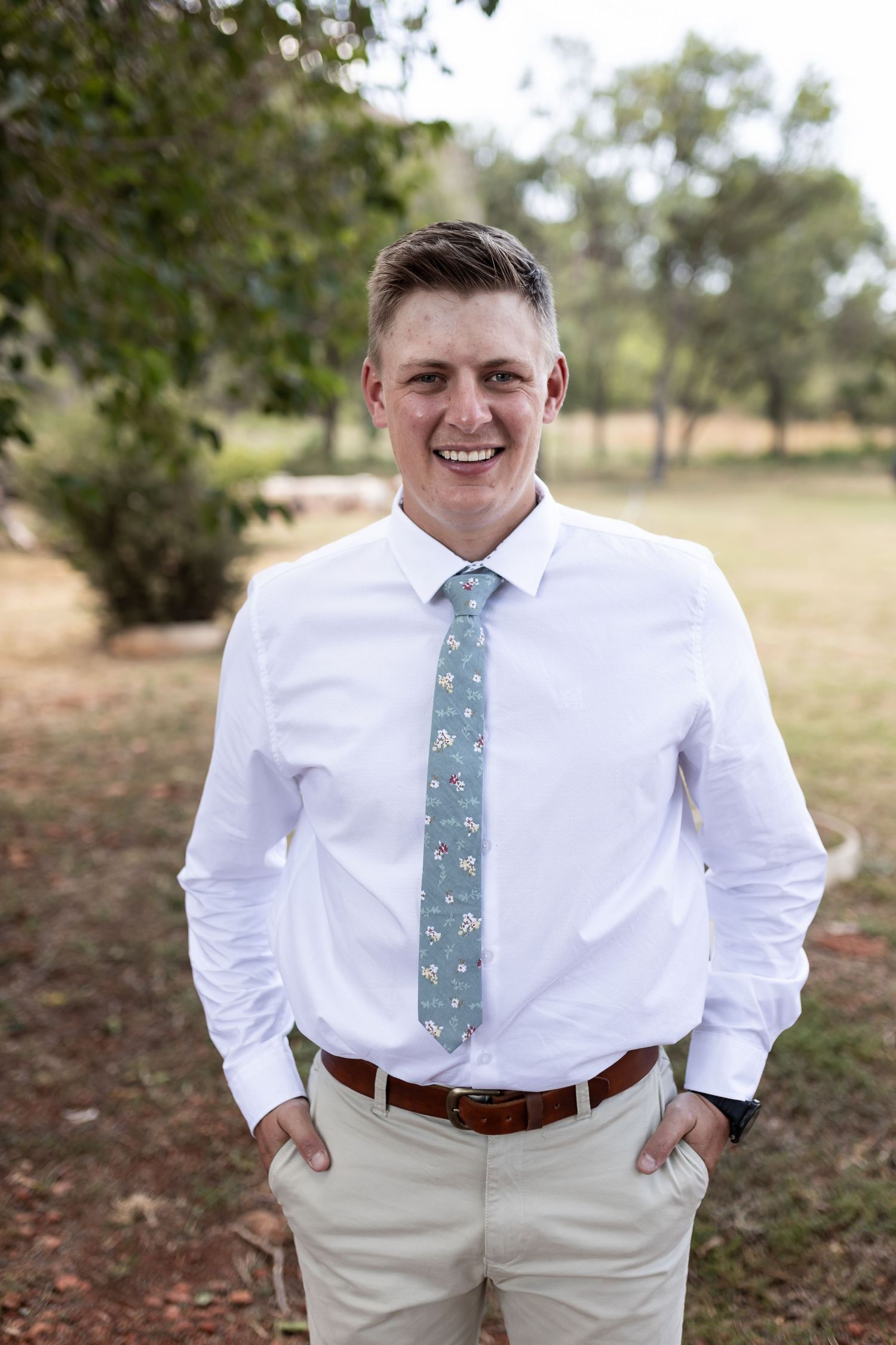 Groomsman wearing light blue patterned tie and white shirt smiles in outdoor setting.