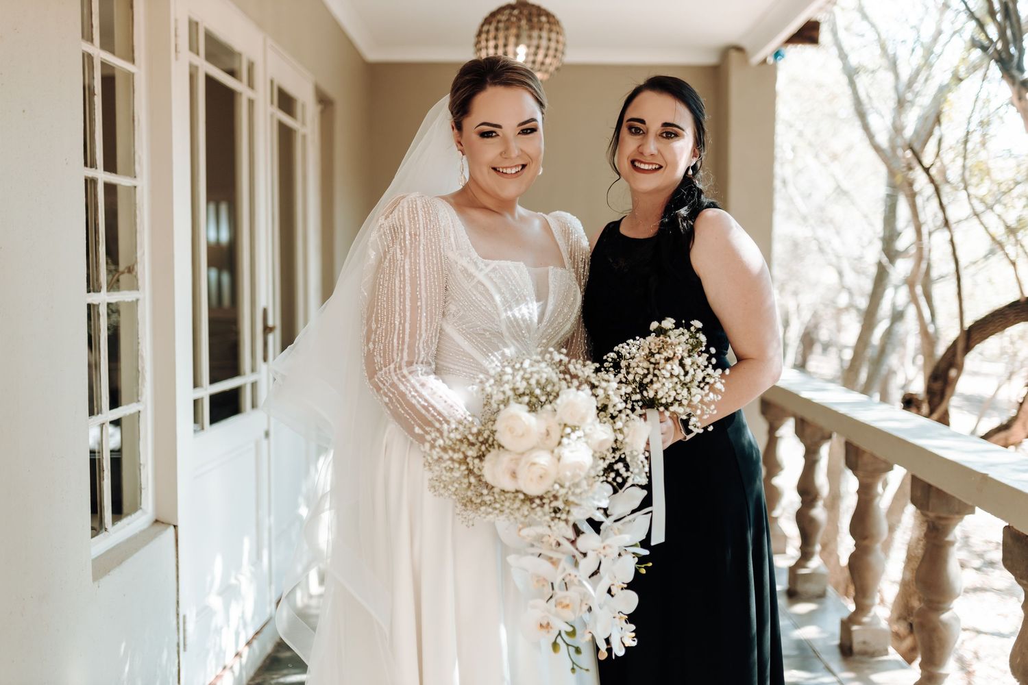 Bride and bridesmaid share a moment on porch while holding white bouquets.