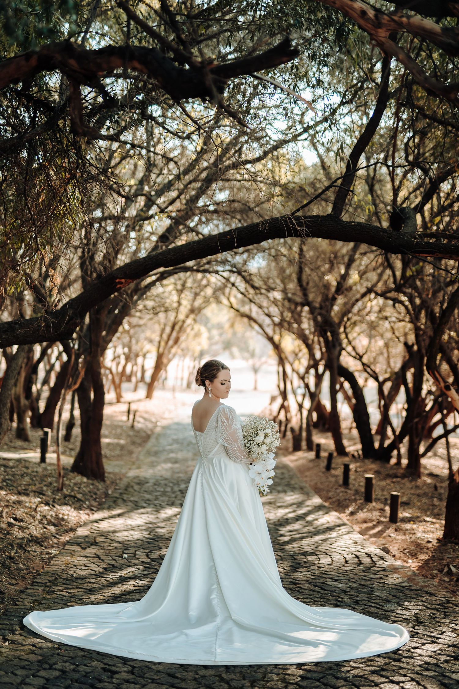 Bride in elegant white gown with long train poses on cobblestone path under tree canopy.