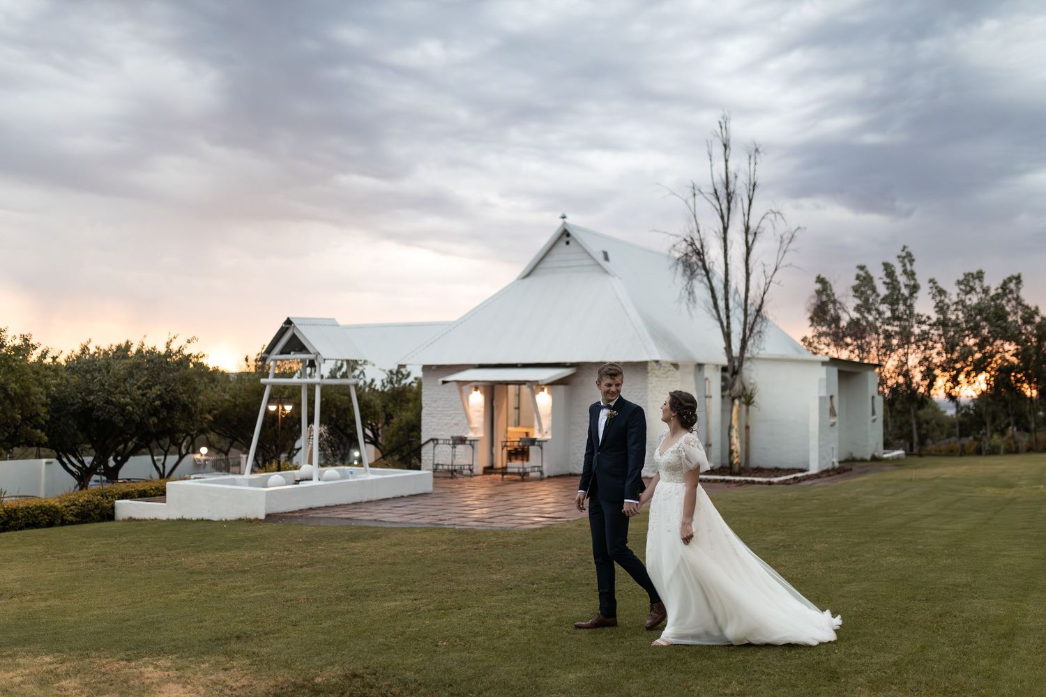 Elegant couple stands on manicured lawn with white chapel venue and dusky sky in background.