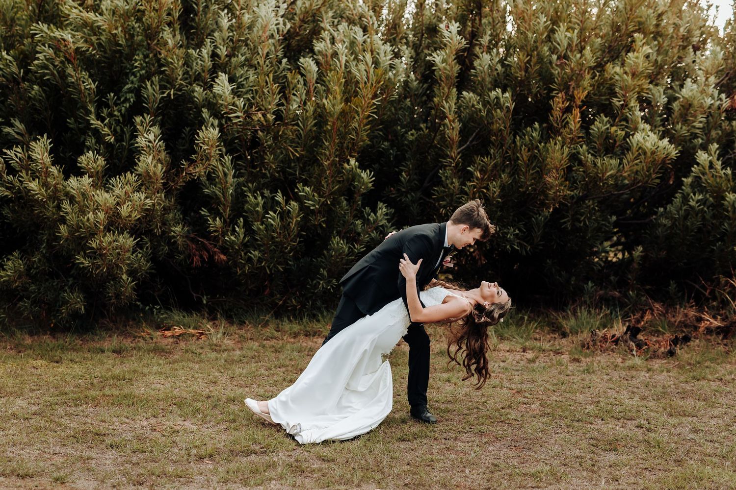 Groom dips bride in romantic pose during outdoor wedding photoshoot.