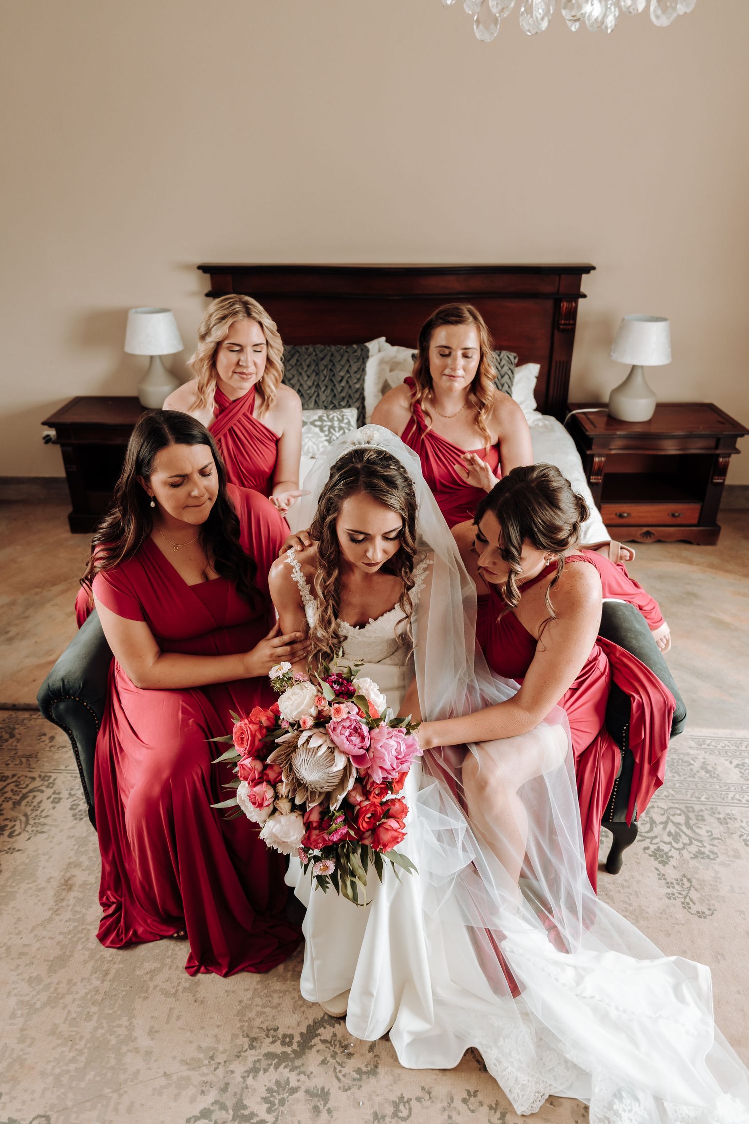 Bridesmaids in red dresses help with final preparations while gathered around a white gown and pink bouquet.