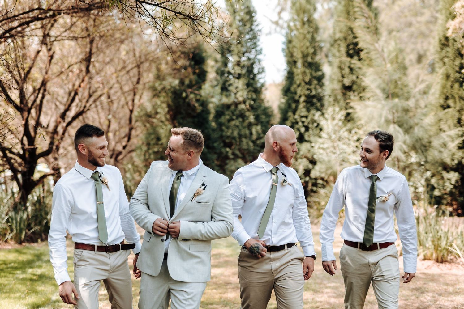 Wedding party in coordinated light attire walks together on outdoor path surrounded by trees.