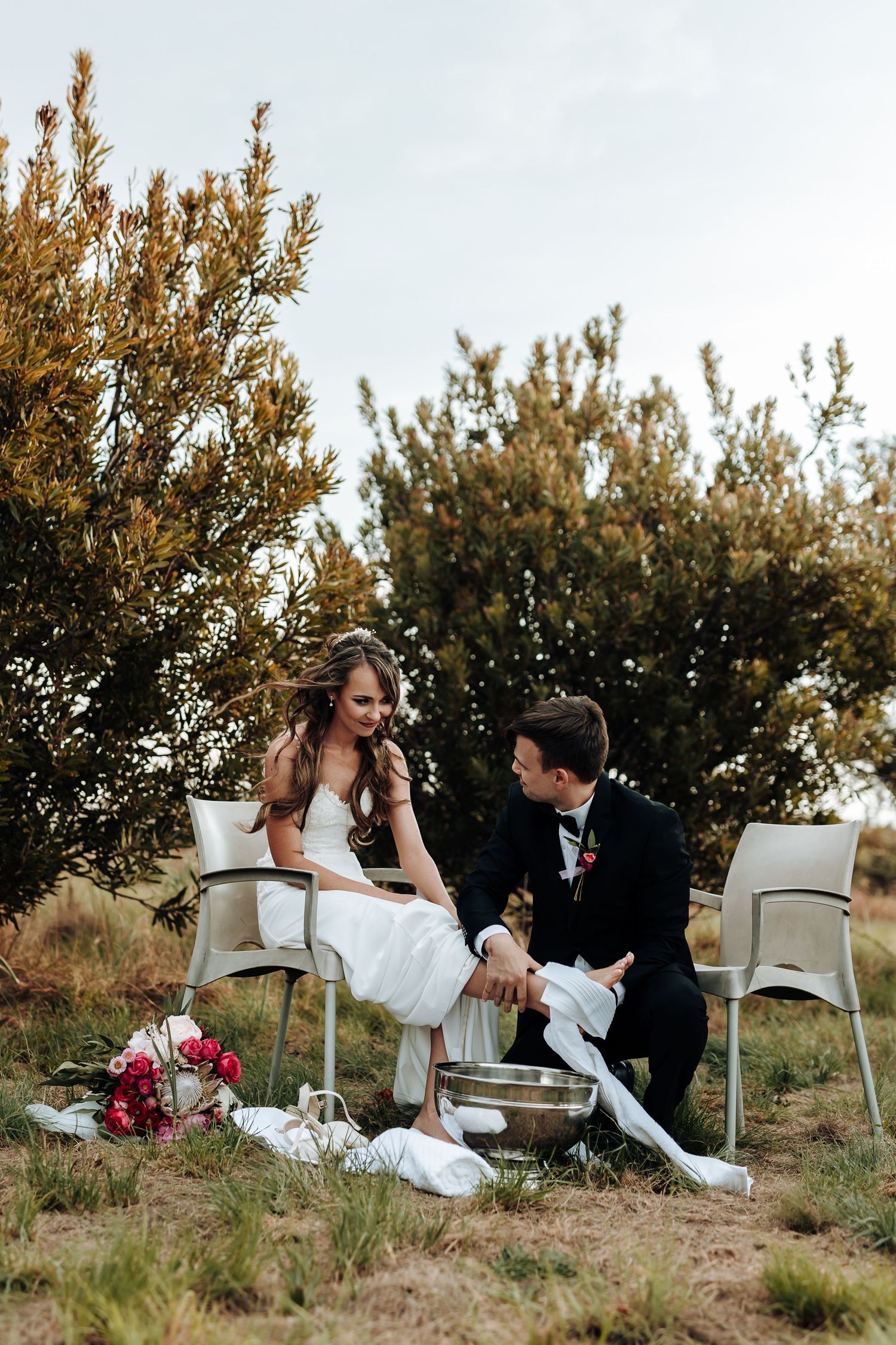 Couple shares playful moment sitting in white chairs during outdoor wedding ceremony.