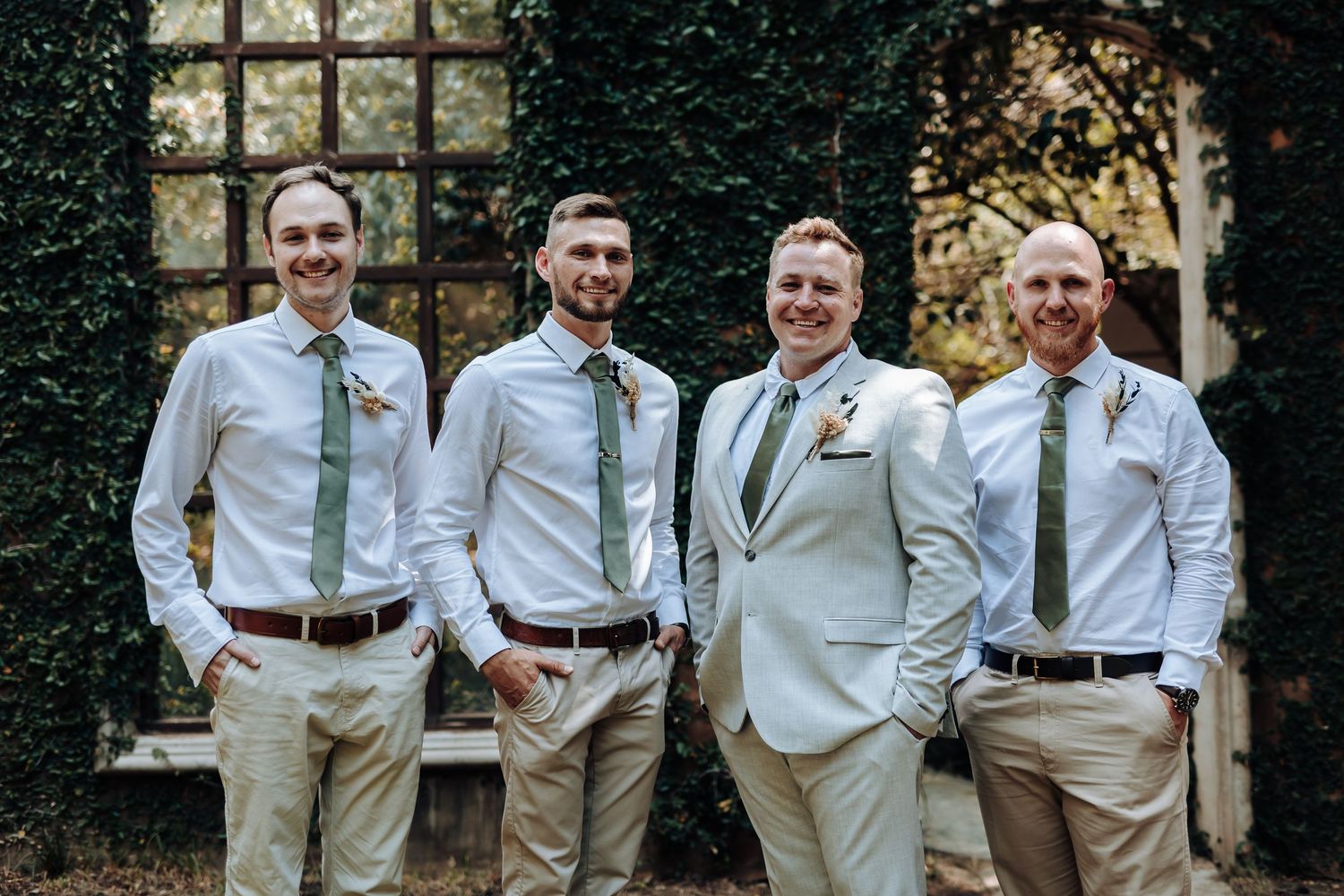 Wedding party in coordinated light colored attire poses together against ivy wall.