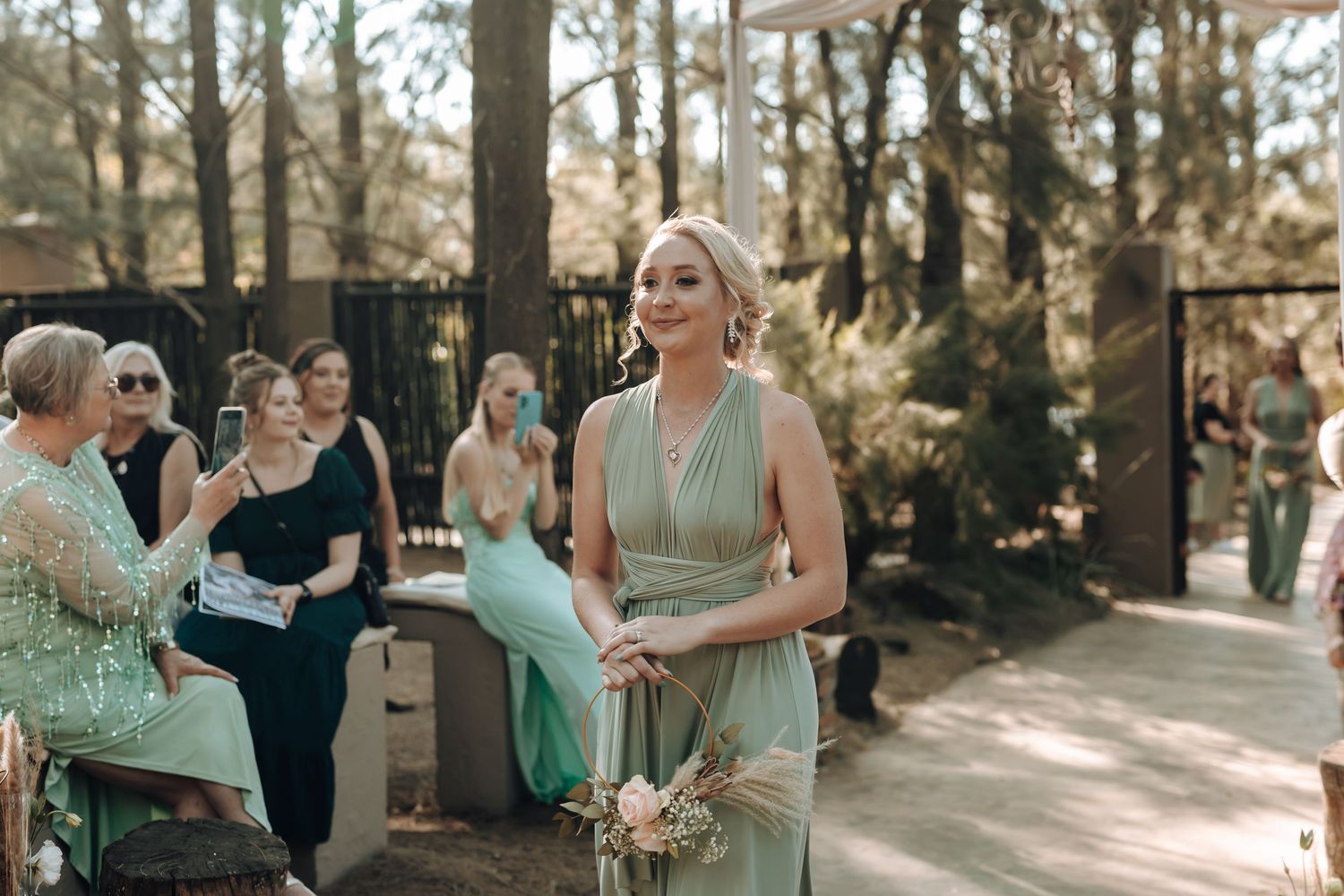 A bridesmaid in a mint green dress holds a bouquet during an outdoor forest wedding ceremony.