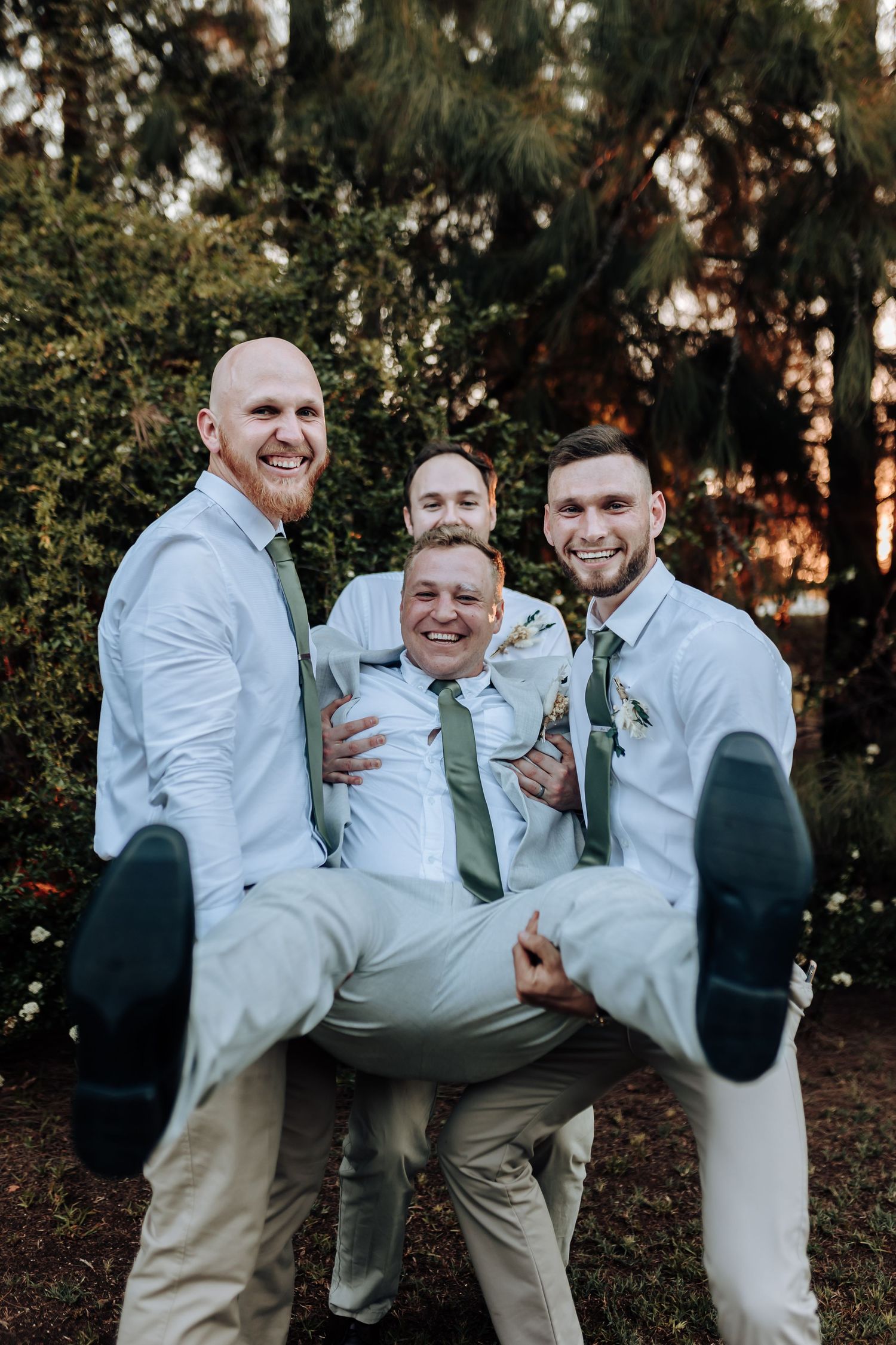 Groomsmen in light colored suits playfully lift fellow wedding party member during outdoor photo session.